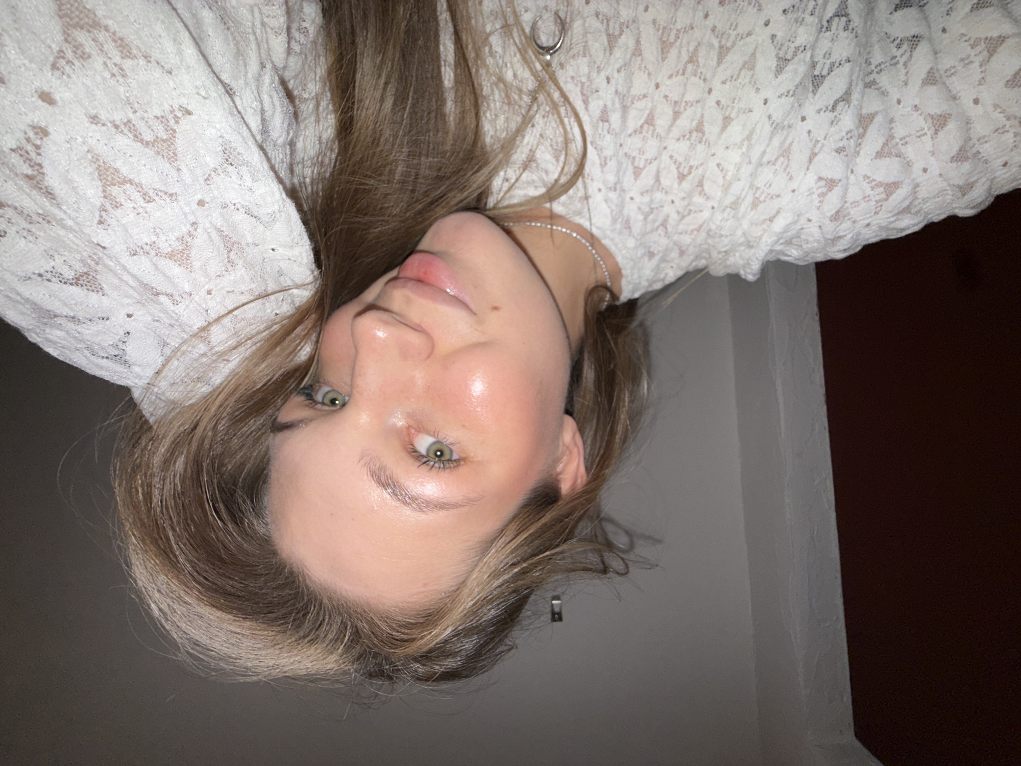 The image shows a close-up, low-angle, indoor shot of a young woman with light brown hair and green eyes. She is wearing a white lace top and a delicate silver necklace with a crescent moon pendant. Her makeup is subtle, highlighting her natural features. The background appears to be a plain wall and a dark floor. There are no visible landmarks or contextual clues to suggest a specific location within Belgrade, Serbia. The scene is static and focuses solely on the subject.