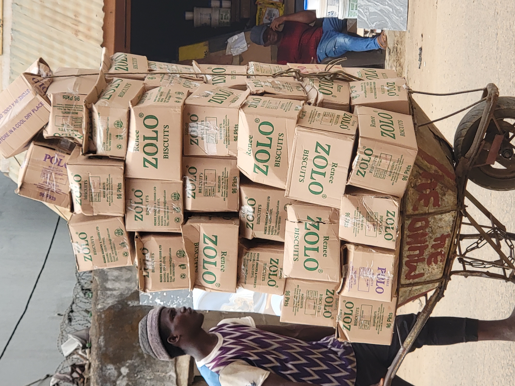 A man pulls a heavily laden wheelbarrow through a street in Maiburiji, Nigeria. The wheelbarrow is piled high with boxes of "Renee ZOLO Biscuits," obscuring much of the barrow itself and its wheel. The man wears a patterned purple and white shirt, dark shorts, a head covering, and is barefoot. He is facing left, looking upward. The ground is paved and dusty. In the background, another man wearing a red shirt and blue jeans is lying down. A shop or building is partially visible behind the first man, with boxes and supplies inside. Barbed wire is visible on a wall.