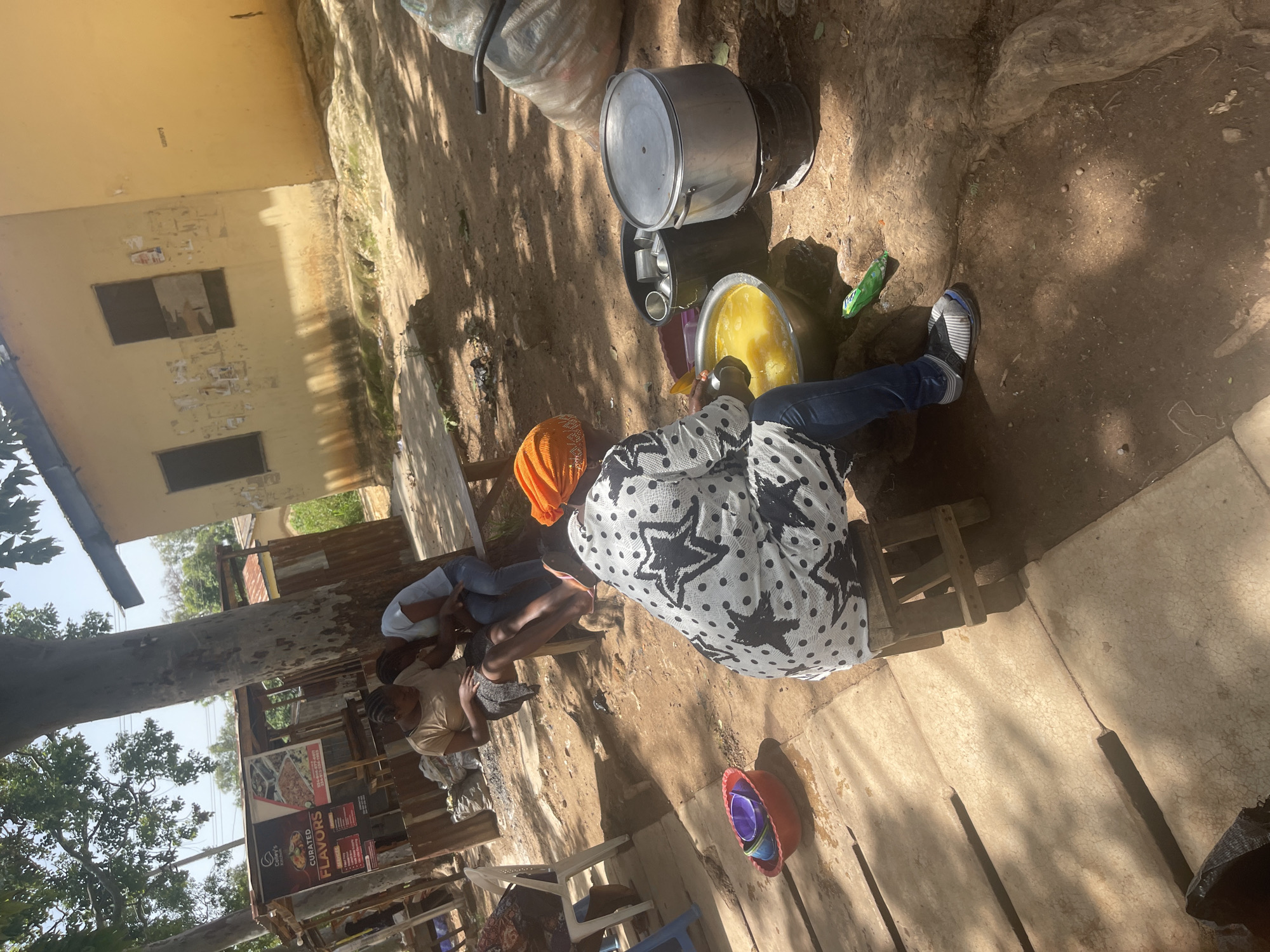 In Rafin Par, Nigeria, on a sunny day, a woman wearing a black and white polka dot patterned shirt and blue jeans is seated on a wooden stool. She is actively stirring a large metal bowl filled with a yellow liquid, likely food preparation. Next to her, two other women are seated on a bench. One is wearing a light brown t-shirt and a dark skirt, while the other is in a light blue t-shirt and jeans, both looking down. In the background, a yellow building with two windows and a corrugated metal structure are visible. The scene is outdoors, with trees providing shade and the ground covered in dirt and some paved sections. Several large cooking pots, a red bowl with smaller bowls inside, and a large bag are also present. The environment suggests a casual, outdoor food preparation or service area.