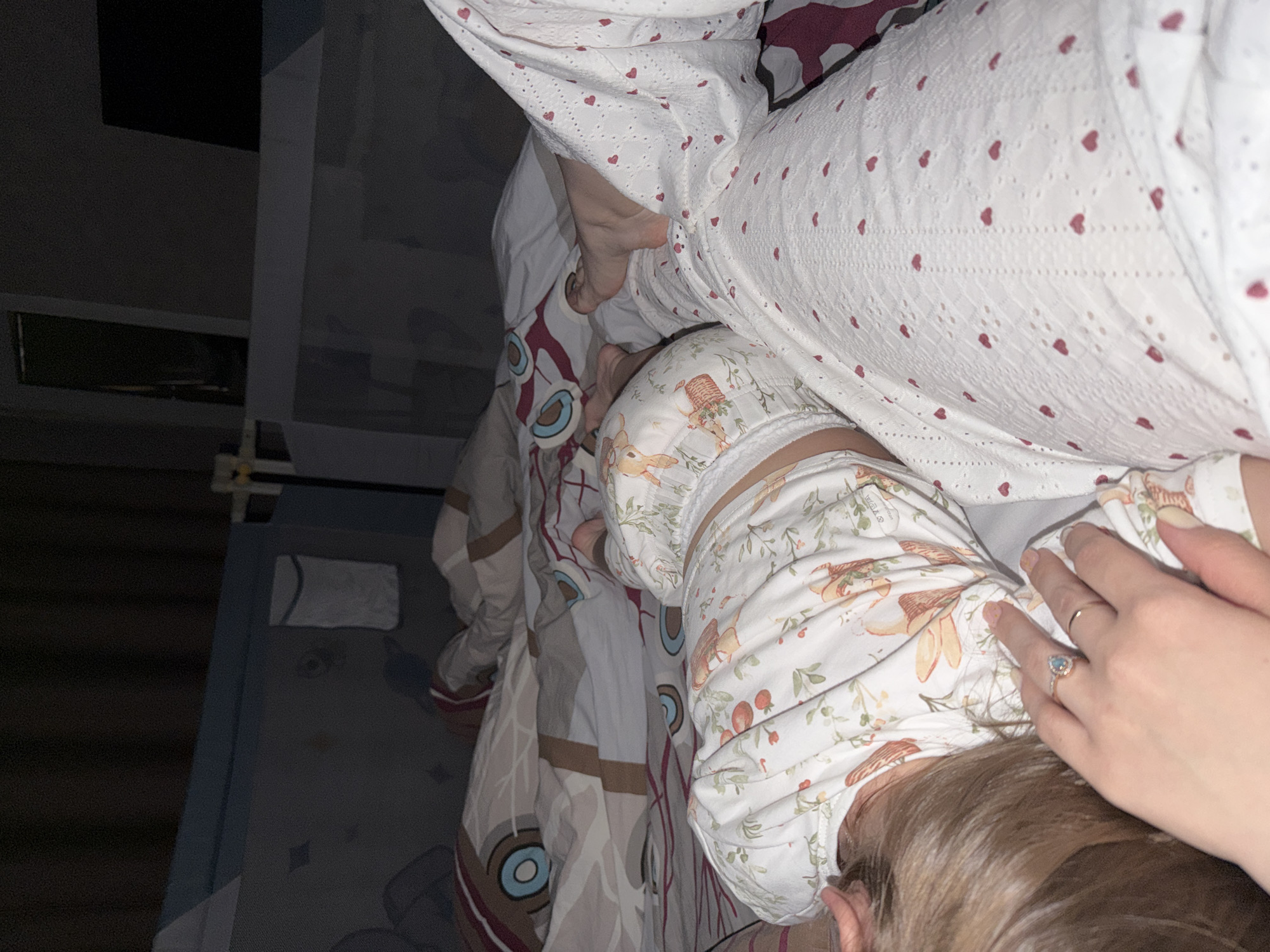 A close-up, overhead shot depicts an adult in white pajamas with red heart patterns lying on a bed next to a small child wearing white pajamas with a pattern of foxes, leaves, and berries. The adult's bare foot is visible near the child. The adult's hand, adorned with a gold band and a ring with a light blue stone, gently rests near the child’s head. The bed is covered in a patterned duvet. The environment appears to be dimly lit. There is no information present in this image to indicate that the location is Reshetnikovo, Russia.