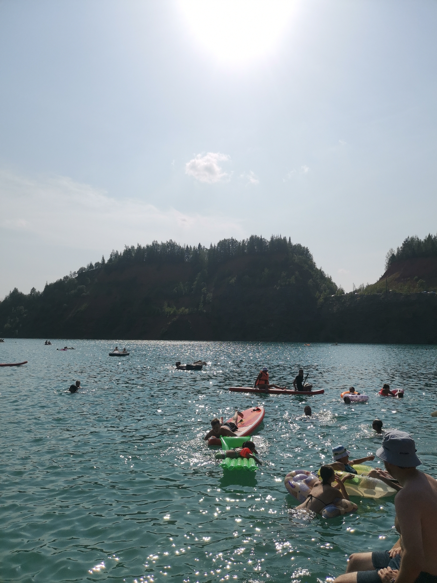 The image depicts a scene at Karyer Izvestnyak, Russia, where a body of water, likely a lake or quarry, is filled with people enjoying various water activities on a sunny day. A steep, tree-covered hill forms the backdrop, with a bright sun visible in the upper right portion of the sky.
In the foreground, several individuals are present. A man wearing a grey bucket hat and dark shorts is seated, facing away from the camera. To his left, a group of people are on inflatable floats. Two children in hats are on a yellow inflatable, and a woman in a black bikini is on a purple and white inflatable. Another person is partially visible on a green inflatable mat.
Further out on the water, a person is on a red paddleboard, accompanied by another individual behind them. Several other paddleboards and inflatable rafts are scattered across the lake. In the middle ground, a group of people are on a raft with an orange ring. In the background, a few individuals are swimming or wading in the water near the shore. The water itself is a vibrant turquoise, reflecting the bright sunlight, creating a shimmering effect. The overall atmosphere suggests a recreational outing on a warm, clear day.