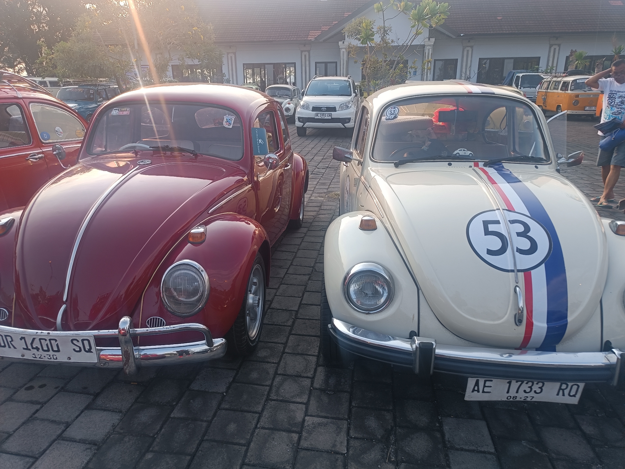 The image shows two classic Volkswagen Beetles parked side-by-side on a paved area in front of a building with a tiled roof. The Beetle on the left is red, and the Beetle on the right is cream-colored with blue and red stripes and the number "53" on its hood. The red Beetle has a license plate that reads "DR 1400 SQ" and "12-30". The cream Beetle has a license plate that reads "AE 1733 RQ" and "08-27". In the background, other vehicles including a white SUV, a white van, and a yellow van are visible. The sun is setting, casting a warm glow over the scene, and trees are present in the background. The overall setting appears to be a parking lot or outdoor gathering space in Kediri, Indonesia.