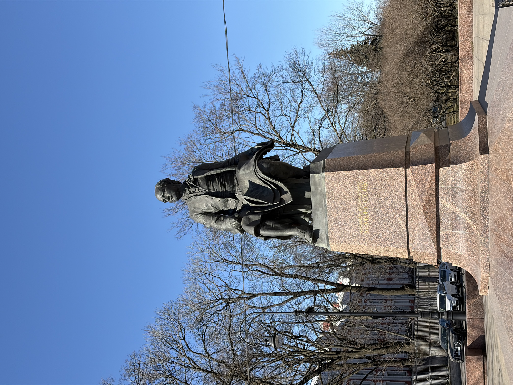 The image depicts a bronze statue of Gavrila Derzhavin, a renowned Russian poet, standing on a granite pedestal. The statue is located in Saint Petersburg, Russia. The background includes bare trees against a clear blue sky, and a glimpse of building facades behind the statue. Several parked cars are visible near the base of the monument.