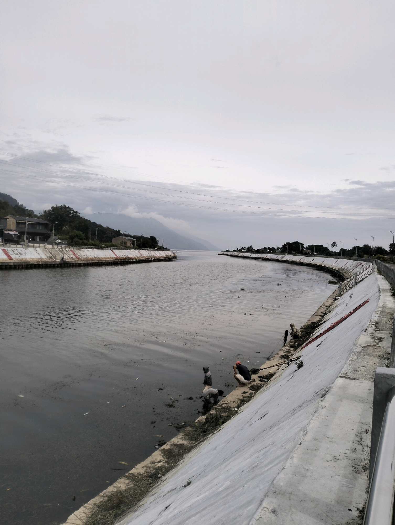 This image depicts the shoreline of a body of water in Pangururan, Indonesia, characterized by a curved, concrete embankment. Several individuals are visible along the embankment and in the shallow water, engaged in what appears to be fishing or some form of water-based activity. The environment is overcast, with a muted color palette dominated by grays and muted greens. Vegetation, including trees and grasses, is present on the landward side of the embankment and on distant hills. Buildings are visible along the left side of the embankment, suggesting a settled area. The water itself appears somewhat murky and contains debris.