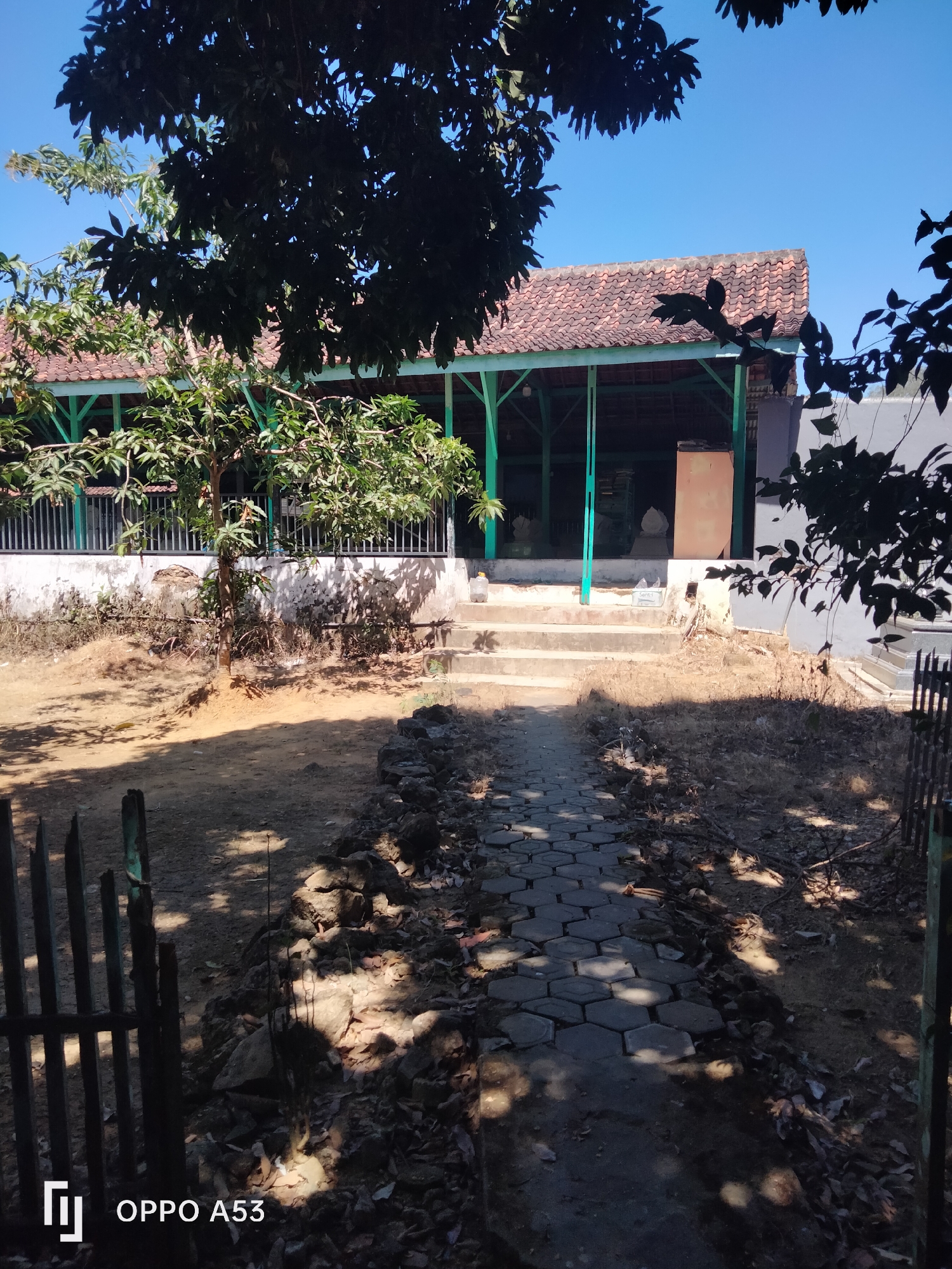 The image depicts an outdoor scene in what appears to be a religious or historical site. In the foreground, a pathway made of hexagonal stone pavers leads towards a building. The pathway is partially shaded by trees. To the left of the pathway, a low, dark fence made of vertical wooden slats is visible. The ground around the pathway is covered with dry leaves, dirt, and some scattered rocks.
The building in the background has a red tile roof and a green-covered veranda supported by green pillars. White walls with some discoloration and peeling paint are visible below the veranda. A metal railing runs along the front of the veranda. Inside the veranda, there are several stone structures, possibly gravestones or religious artifacts, and a tall, rectangular, brown object. There are also some trees and foliage growing around the building. The sky above is a clear, bright blue.
The bottom left corner of the image includes a white logo and the text "OPPO A53," indicating the device used to capture the photograph.
Without additional information or context, it is not possible to definitively confirm the location as the city of Badung, Indonesia. However, the architectural style and vegetation could be consistent with a location in Southeast Asia.
