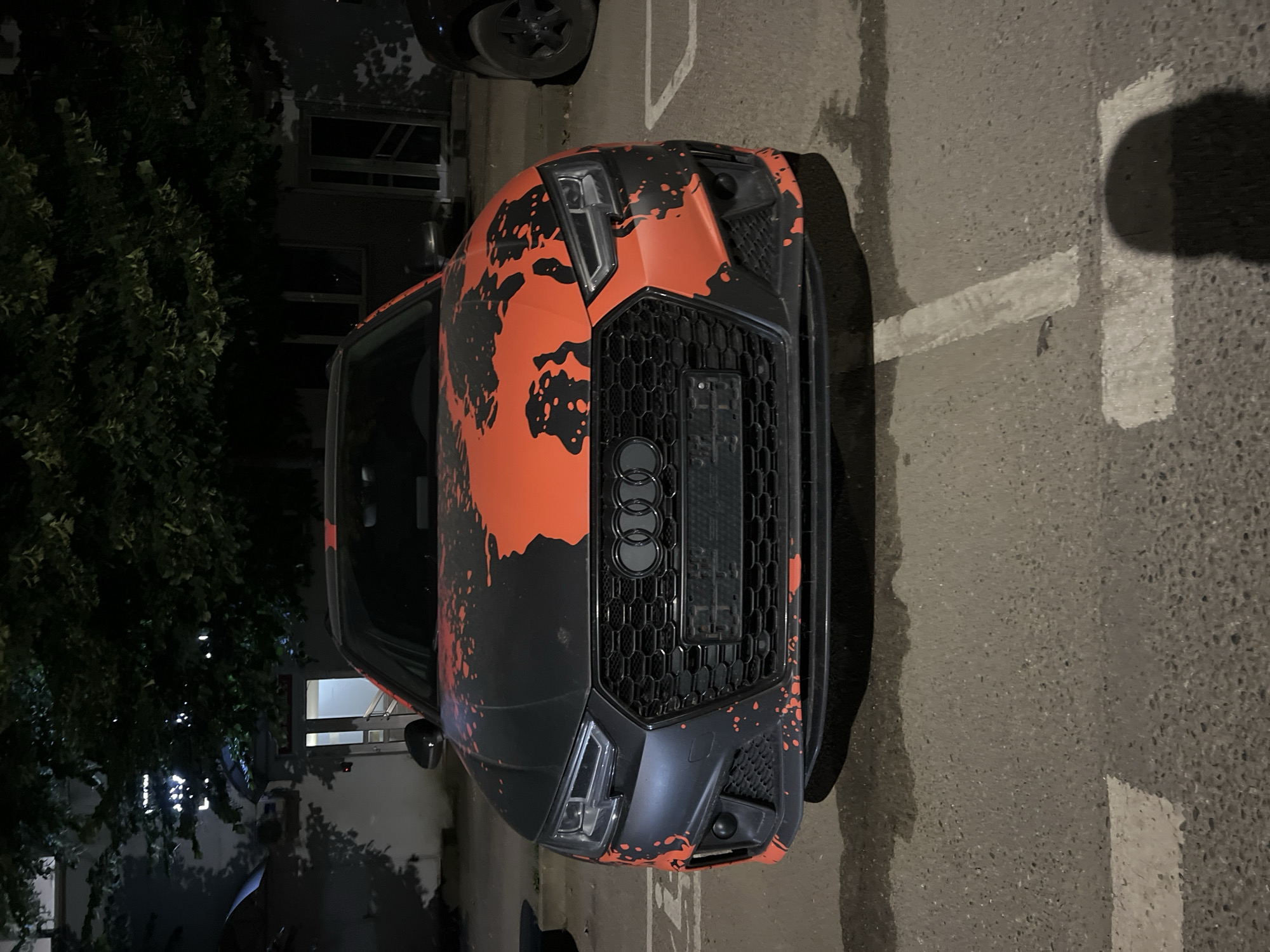 The image displays a front view of a black Audi car with an orange and black abstract splatter design. The car is parked on an asphalt surface marked with white lines, suggesting a parking lot. The car's headlights and grille are clearly visible, featuring the distinctive Audi rings logo. The scene is illuminated by artificial light, indicating it is nighttime. To the left of the car, there is a dense green tree, and a building with windows can be seen in the background. The overall atmosphere is dark, with shadows cast by the car and surrounding elements. No people are visible in the image. The location context is Krasnodar, Russia.