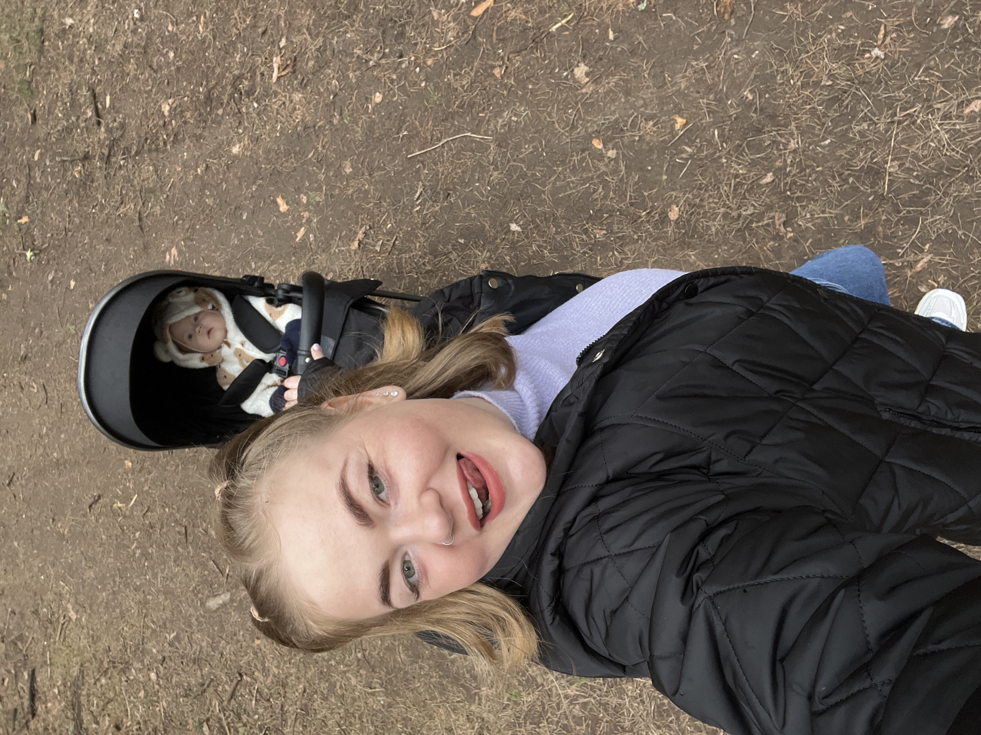 An outdoor, high-angle, selfie-style photograph shows a young woman and an infant in a stroller on a dirt path. The image is rotated, with the subjects appearing diagonally.
The young woman, positioned prominently in the foreground, is smiling broadly with her tongue slightly out, looking directly at the camera. She has light brown or blonde hair, pulled back in some sections, and visible ear piercings and a septum nose ring. She is wearing a black quilted jacket over a light purple/lavender sweater and blue jeans, with a white sneaker visible on her right foot.
Behind and to the left of the woman, an infant is visible lying in a dark-colored stroller. The baby is dressed in a cream-colored, possibly bear-themed, hooded outfit with ears and small mittens, looking calmly upwards.
The background consists of a natural ground surface covered with dry, reddish-brown dirt, scattered with fallen leaves and small twigs, characteristic of a park or woodland path. The lighting suggests it is daytime. The overall mood of the image is cheerful and affectionate, capturing a moment between a caregiver and a baby. There is no visible text in the image. The setting is consistent with an outdoor area in Chaikovsky, Russia.