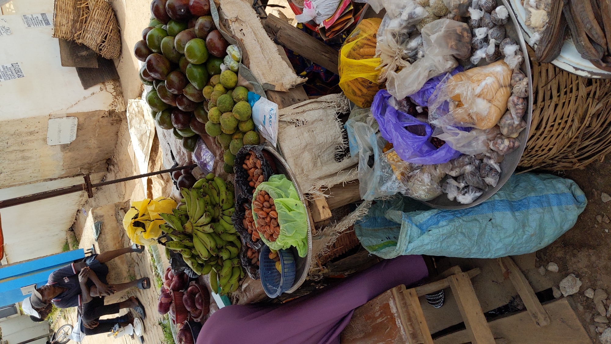 The image captures a bustling outdoor market scene, likely in Ado, Nigeria, characterized by an array of fresh produce and packaged goods displayed on makeshift stalls and the ground.
In the upper portion of the image, a large pile of green and reddish-brown avocados or large mangoes is prominently displayed. Below them, numerous green citrus fruits, possibly limes or small oranges, are visible. To the left, several bunches of green bananas are laid out. The center features a metal tray holding various nuts or seeds, some in black plastic and others in a light green plastic bag, resembling kola nuts or similar local produce. Another large metal basin or tray on the right side contains a variety of goods packaged in clear, purple, and yellow plastic bags, including what appear to be dried spices, grains, or snacks. A large woven basket to the far right holds dark, elongated dried items.
On the left side of the frame, a man wearing a dark t-shirt, shorts, a white cap, and blue flip-flops is seated, leaning forward with his hands on his lap, seemingly resting or waiting. A vendor, partially visible in the lower center, is clad in a purple garment.
The ground is rough, composed of dirt and gravel, indicative of an informal market setting. Various objects like more woven baskets, a light blue woven sack, and wooden planks contributing to the stalls are visible. The bright daylight suggests the time of day is morning or afternoon.
Visible text elements include: "BCK010070", "1910603072", "ALUQUON", and "1706189649" written on light-colored surfaces. Another inscription mentions services like "CLEAN WORK", "HAIR PRESS", and "CLEANERS". A package, possibly for a face mask, is labeled "K-MASK". The overall scene portrays a typical vibrant, busy market environment.