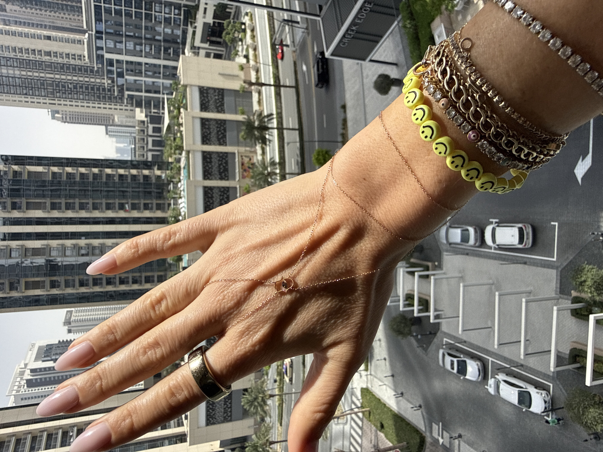 The image shows a fair-skinned person's hand and wrist adorned with various pieces of jewelry against an urban backdrop. The hand is angled, fingers extended, and manicured with light pink polish. On the fingers is a gold band. A delicate gold chain with a central design drapes across the hand. The wrist is stacked with bracelets, including a tennis bracelet, multiple gold chain bracelets, and one made of yellow smiley face beads.
The background reveals a cityscape, including modern high-rise buildings, palm trees, a road with moving cars, and parking areas with parked cars. A sign reading "CREEK EDGE" is also visible. This indicates the location is likely in the Creek Edge area of Dubai, United Arab Emirates. The lighting suggests it is daytime. There are no other people or interactions directly visible in the image.