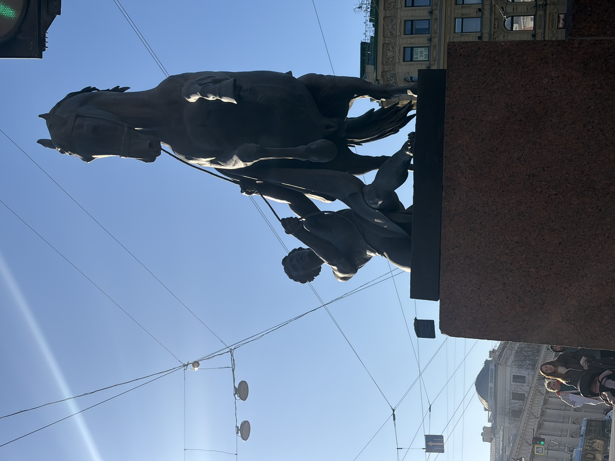 Here's a description of the image:
The image shows a low-angle view of the "Horse Tamers" sculpture on Anichkov Bridge in Saint Petersburg, Russia. The sculpture depicts a man struggling to control a rearing horse. The sculpture is mounted on a reddish-brown granite pedestal. In the background, there is a blue sky with some electrical wires running across it, and buildings with architectural details. At the bottom of the image, blurry pedestrians are visible, and an adjacent building's ornate facade can be seen. A traffic light is visible.