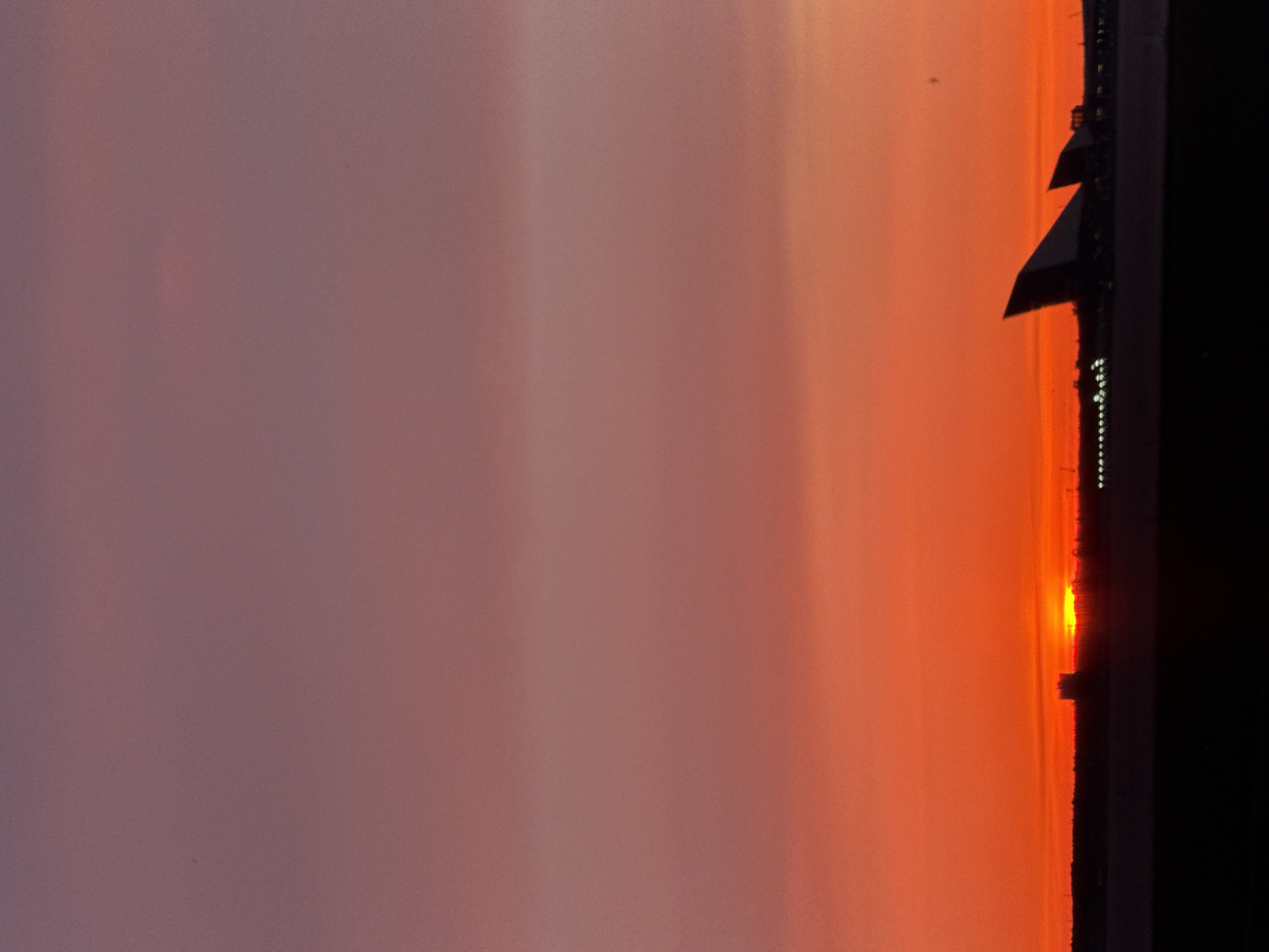 The image depicts a sunset over Saint Petersburg, Russia. The sky displays a gradient of colors, transitioning from a soft purple at the top to a vibrant orange and red near the horizon. Silhouetted against the sky are the distinctive sail-like structures of the Lahta Center. The foreground is dark and featureless, suggesting it is nightfall or a heavily shadowed area. The horizon is marked by a line of dark structures and distant lights. The overall impression is a dramatic and atmospheric view of the city at dusk.