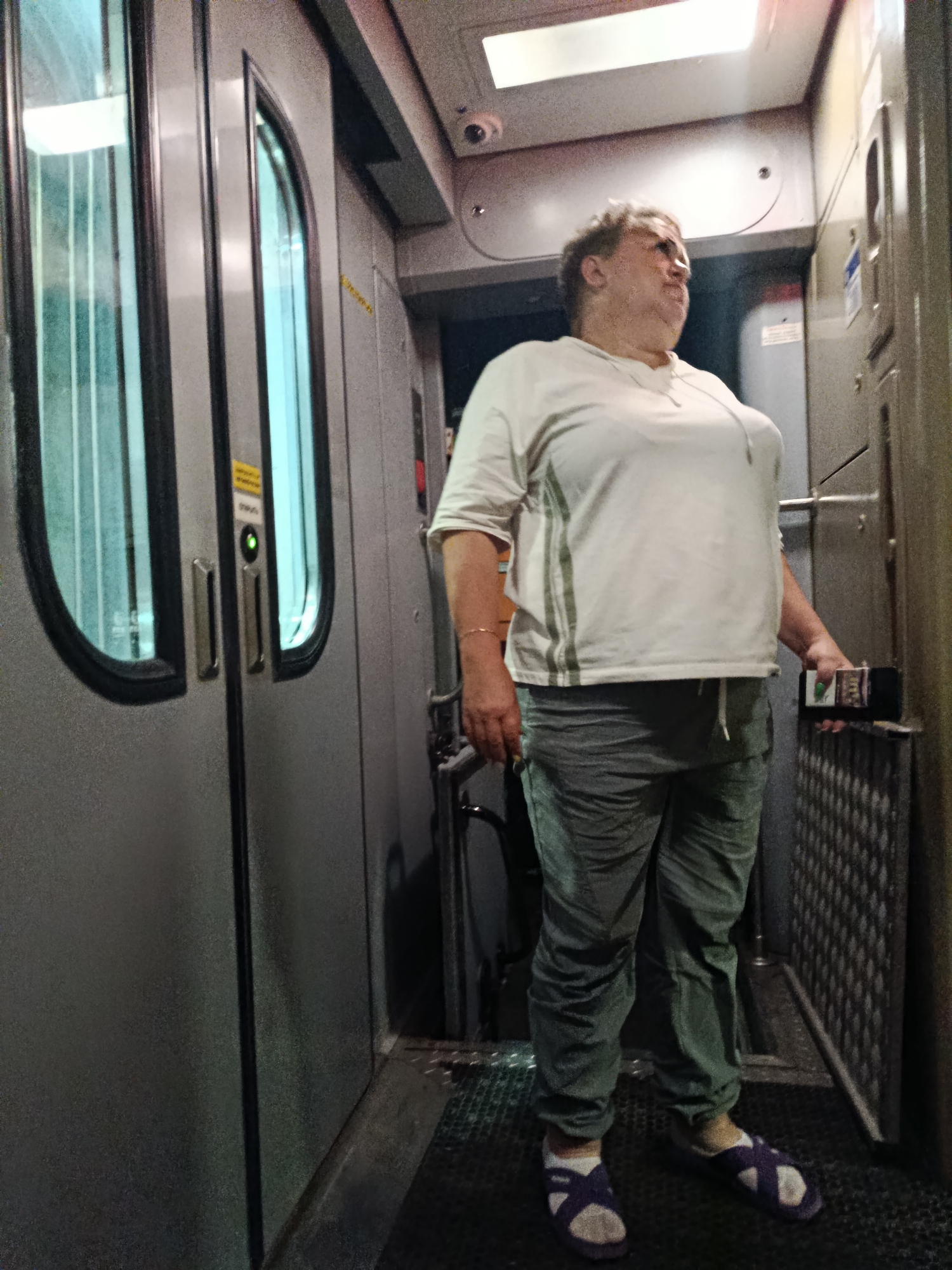 A person stands in a narrow corridor. The person is wearing a white t-shirt with green stripes on the sleeves and grey pants. They are also wearing purple and white sandals. The person is holding a black device with a green and white label in their right hand. The corridor has double doors on the left with tinted windows and metal handles. There is a metal grate on the floor. The walls are grey and there is a light on the ceiling. The environment appears to be a train or bus interior. There is no indication of the location being in Rtishchevo, Russia.