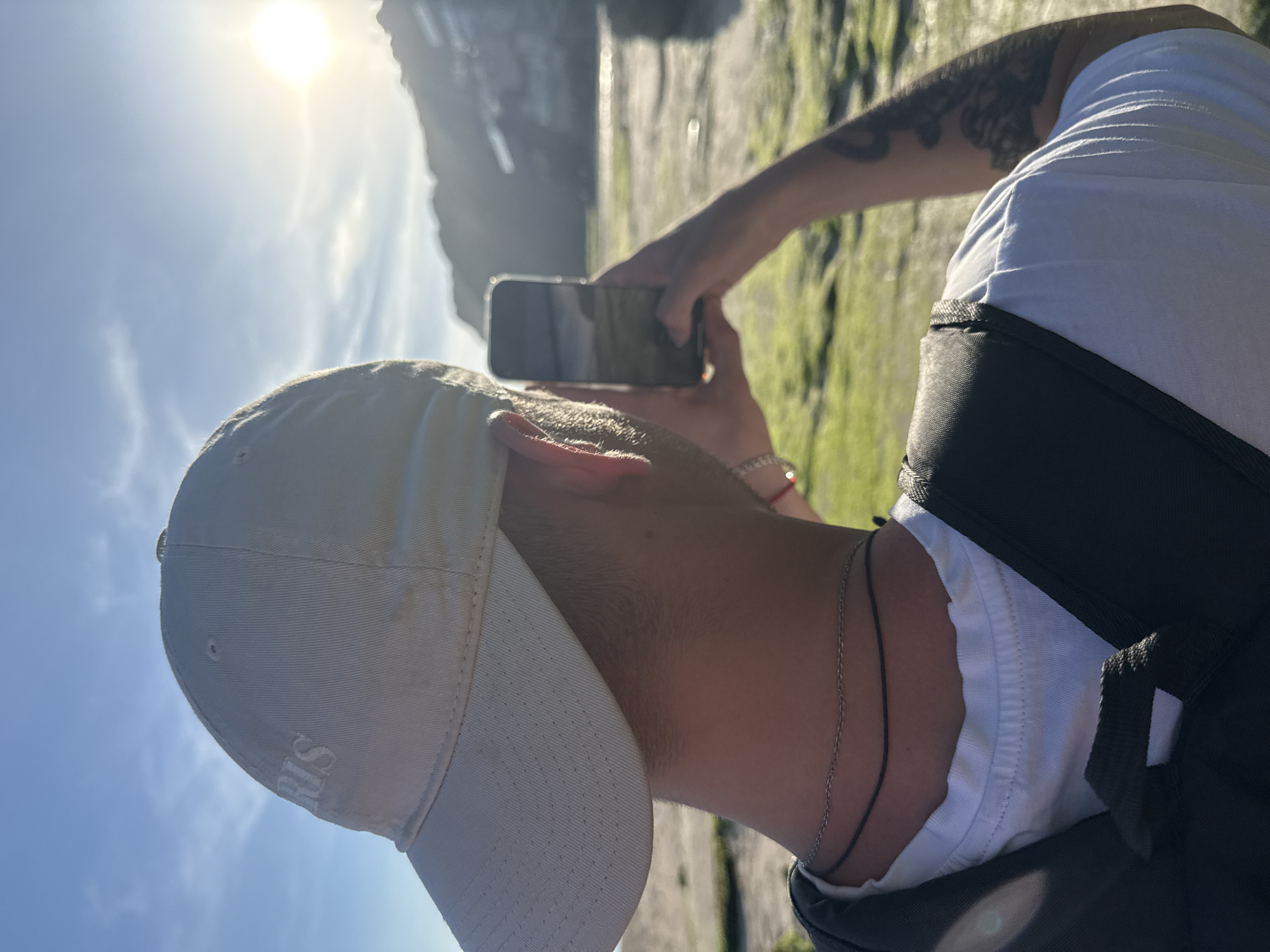 A person wearing a white t-shirt, a black backpack, and a white baseball cap is holding a smartphone and taking a picture. The background appears to be a coastal area with green vegetation and a rock formation in the distance. The sun is shining brightly in the sky. The location is likely the coast of Kuta Selatan, Indonesia.