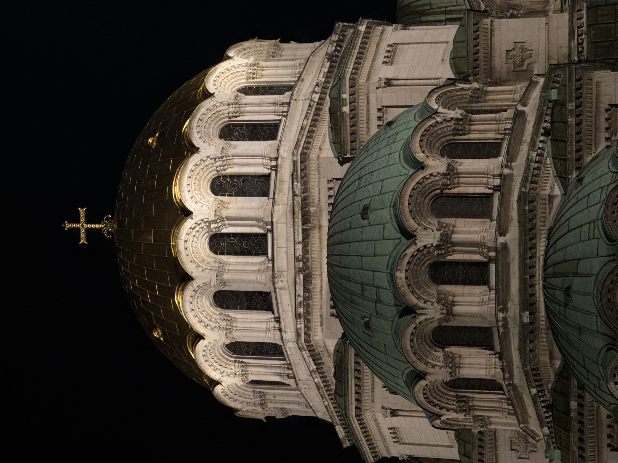 The image shows the Alexander Nevsky Cathedral in Sofia, Bulgaria, at night. The golden dome with a cross is on the left, contrasted against the dark sky. To the right of the golden dome are the cathedral's architectural details, including arched windows and a green dome. The building's facade is illuminated, highlighting its intricate details. The image is oriented vertically, with the sky taking up much of the left side.