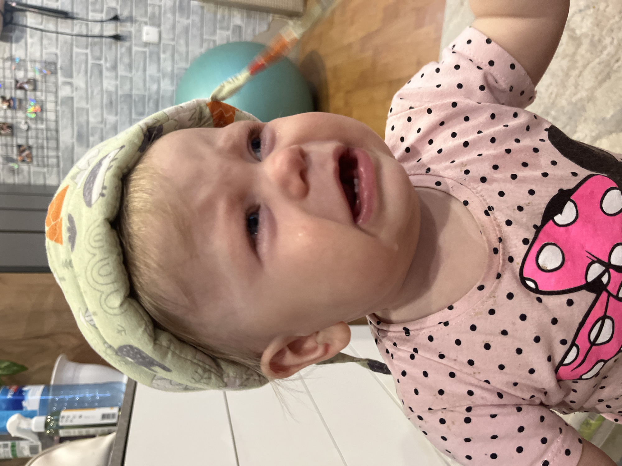A close-up shot depicts a crying toddler. The child is wearing a light green padded head covering and a pink, short-sleeved shirt with black polka dots and a Minnie Mouse bow design. A tear is visible on the child's face. In the blurred background, there's a light green exercise ball and what appears to be a white countertop with bottles. The location is Vesyoloye, Russia.
