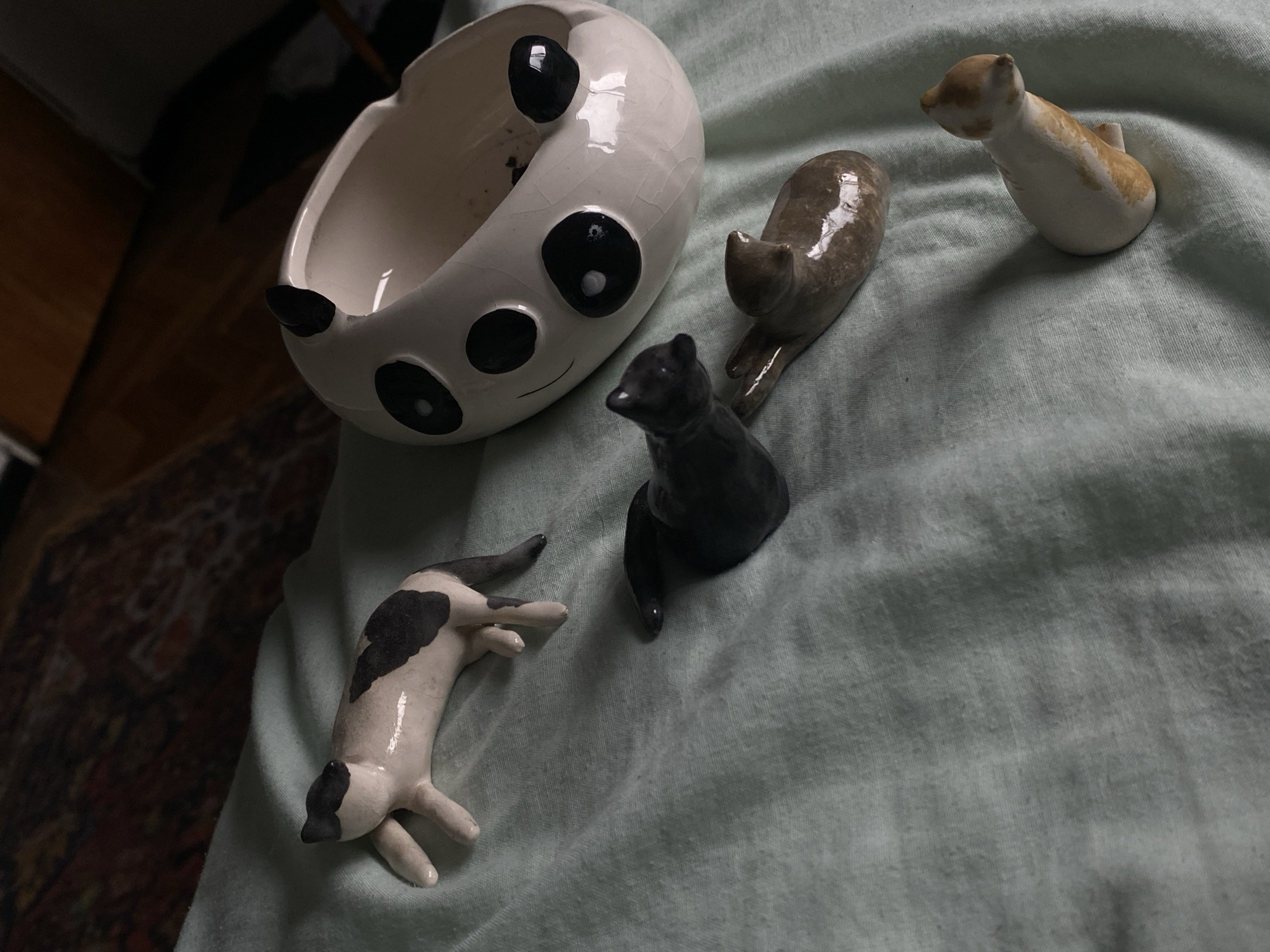 The image shows a collection of small ceramic objects on a light blue fabric surface. The objects consist of a white ceramic bowl with black markings resembling a panda face, and four small ceramic animal figurines: a black bear, a brown cat-like creature, a white and brown cat-like creature with a small upright tail, and a white and black reclining cat-like creature. The fabric surface appears to be slightly wrinkled and has a subtle texture. The background shows a section of a dark wooden floor. The location is not discernible.