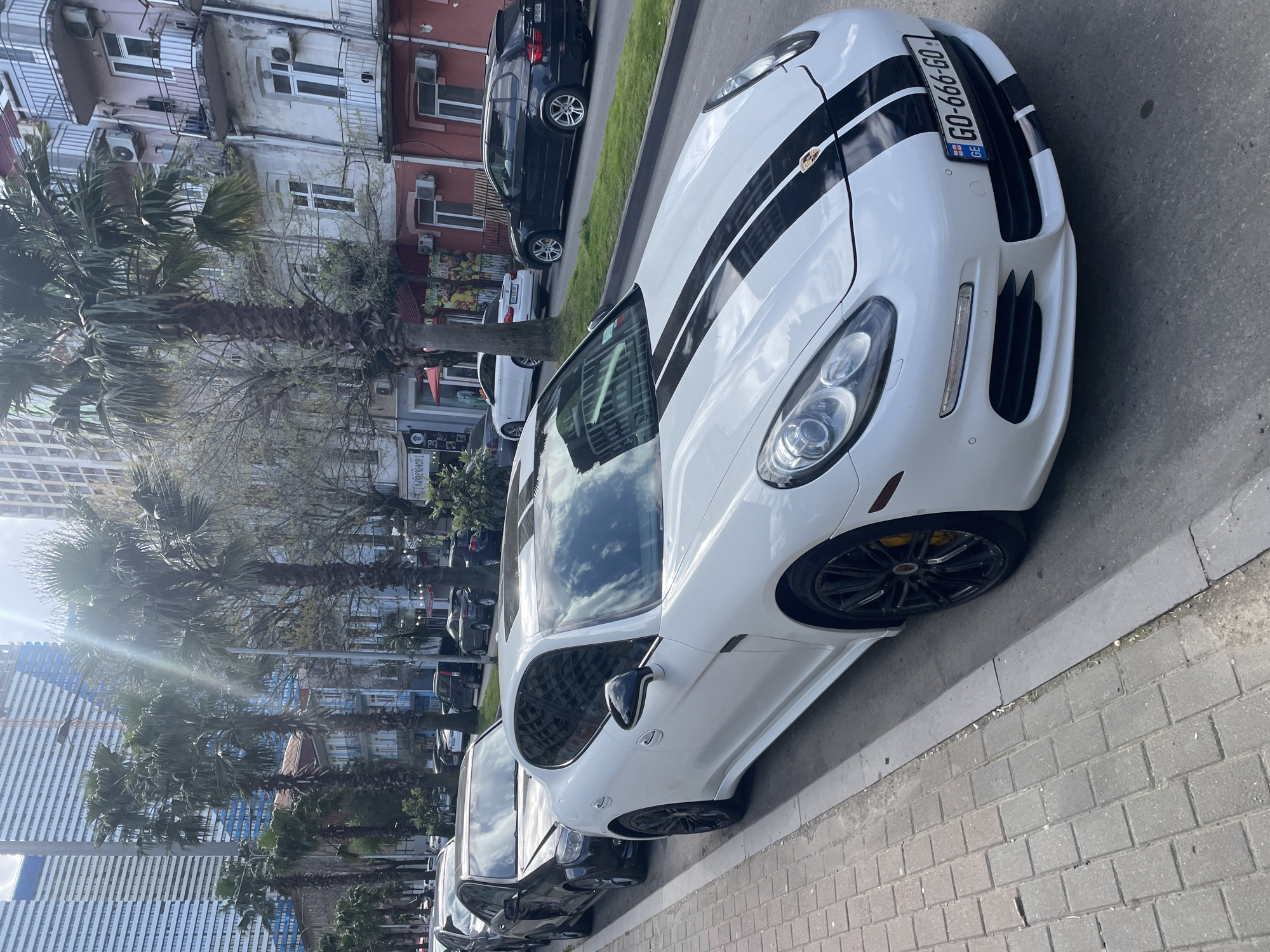 The image depicts a street scene in Batumi, Georgia. Dominating the foreground is a white Porsche Panamera with black racing stripes and a Georgian license plate reading "GO-666-GO". The car is parked along a brick-paved sidewalk. A line of other parked cars, mostly dark-colored sedans and SUVs, extends further down the street. On the left side of the image, palm trees line the sidewalk, providing a tropical feel. Behind the palm trees, tall modern buildings, likely residential or commercial, are visible. On the right side of the image, older, low-rise buildings with a mix of architectural styles and colors can be seen. The sky is partly cloudy, and the sun is shining. There are no visible people in the immediate vicinity of the Porsche, but there are some people visible in the background.