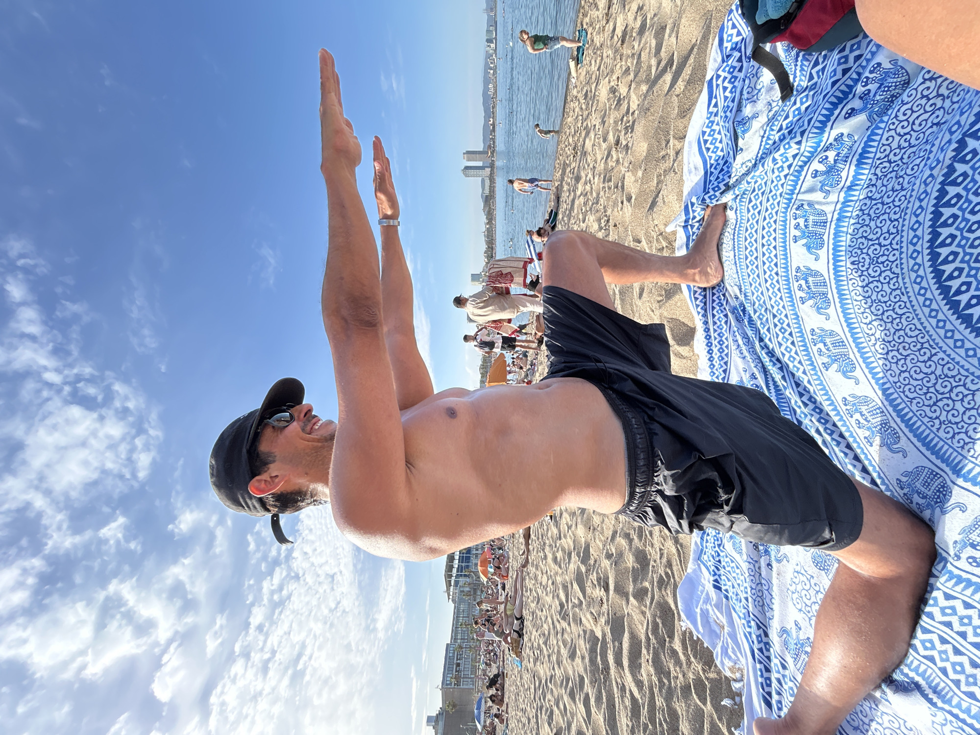 A shirtless man wearing a black baseball cap, sunglasses, and black swim shorts is performing a yoga pose on a sandy beach in Barcelona, Spain. He is kneeling on a blue and white patterned towel with his left leg forward and his right leg bent behind him. His arms are extended forward, parallel to the ground, with his palms facing down. The man is smiling and looking to his right.
In the background, there are many other people on the beach, some sunbathing and others walking near the water. Beach umbrellas are visible in various colors. The sea is calm, and there are buildings and two distinctive twin towers in the distance, likely the W Barcelona Hotel and the Mapfre Tower. The sky is blue with scattered white clouds, indicating a sunny day. The overall scene captures a recreational moment on a crowded beach.