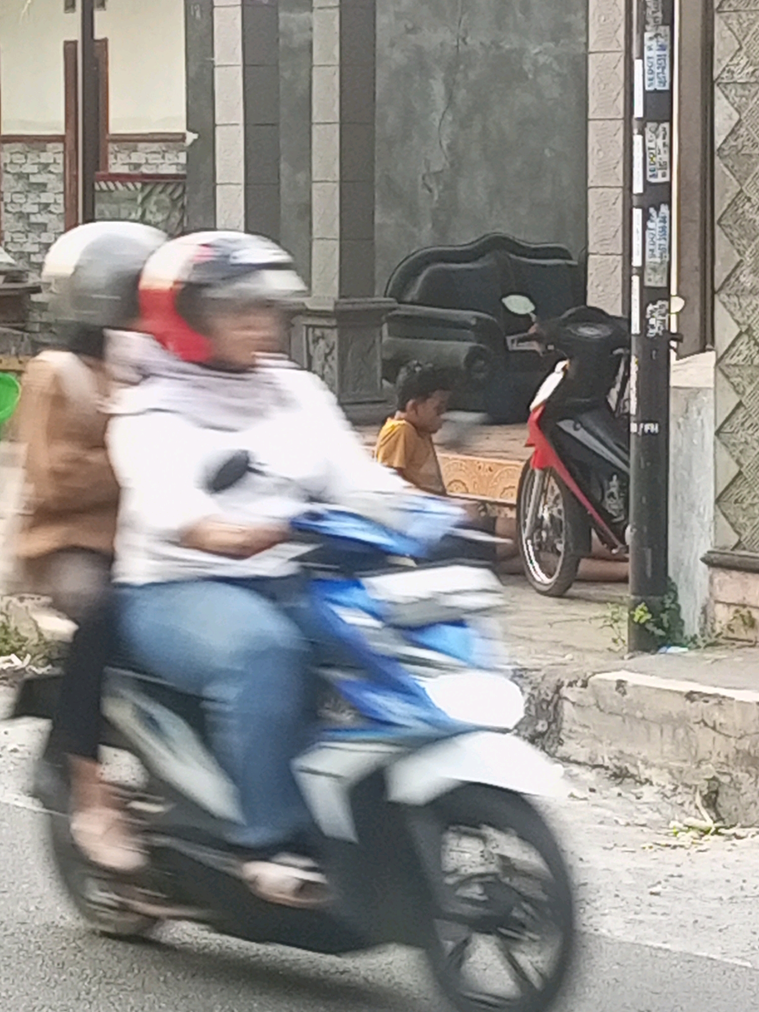 Two individuals, a driver and a passenger, are riding on a blue and white motorcycle, moving from left to right. The driver, in front, wears a helmet with red accents and a white head covering, along with a white top and blue jeans. The passenger, behind, wears a grey helmet and a brown jacket. The motorcycle is in motion, evidenced by the motion blur on the figures and the vehicle. In the background, a young boy in a yellow shirt is seated, looking to the left. A parked red and black motorcycle is visible to the right of the boy, with a black stand. A utility pole with stickers is positioned to the right of the parked motorcycle. The scene appears to be a street in Ngasem, Indonesia, with concrete structures and some vegetation present.