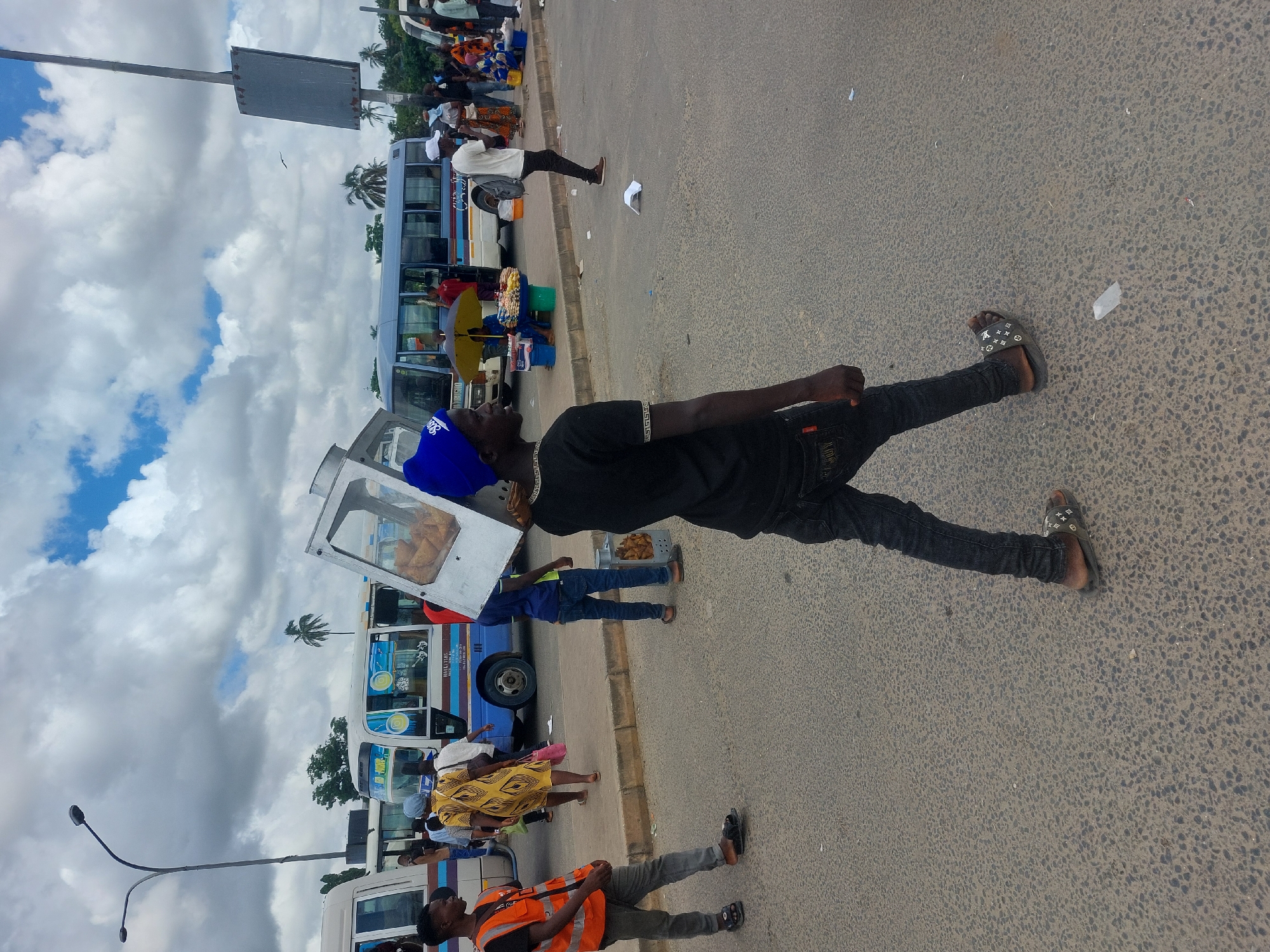 The image depicts a street scene in Dar es-Salaam, Tanzania. Several white minibuses are parked along the side of a road, with people standing around them. Some individuals appear to be selling snacks from a glass display case mounted on one of the buses. The road is paved and leads into the distance. The sky is partially cloudy. A man in black clothing and sandals stands on the road, facing the direction of the parked buses. Other individuals are visible further down the road, including someone wearing a bright orange vest. A light pole and a sign are visible on the left side of the image.