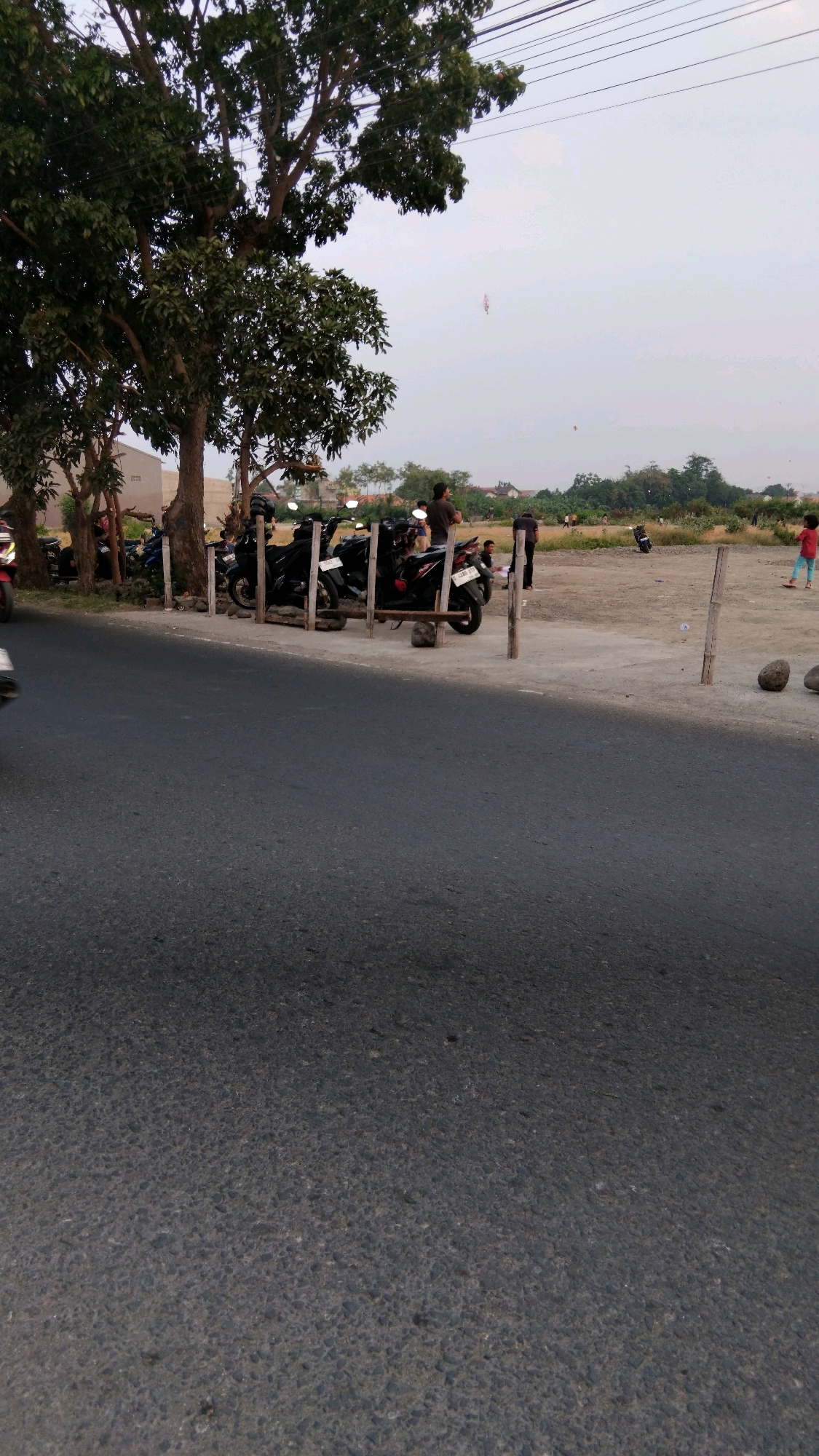 The image depicts an outdoor scene near a road in Tegal, Indonesia, likely in the late afternoon based on the lighting. In the foreground, a dark asphalt road occupies the bottom half of the frame. To the left and slightly further back, a row of parked motorcycles is visible, secured by wooden posts. Beyond the motorcycles, a large tree with dense foliage stands prominently, casting shadows. In the mid-ground, an open, unpaved lot is present. Several people are scattered across this area. To the right, a young child in a red shirt and blue pants is visible. Further in the background, a line of trees and buildings suggests the presence of a residential or semi-urban area. The sky is overcast and light. Two kites are visible flying in the sky.