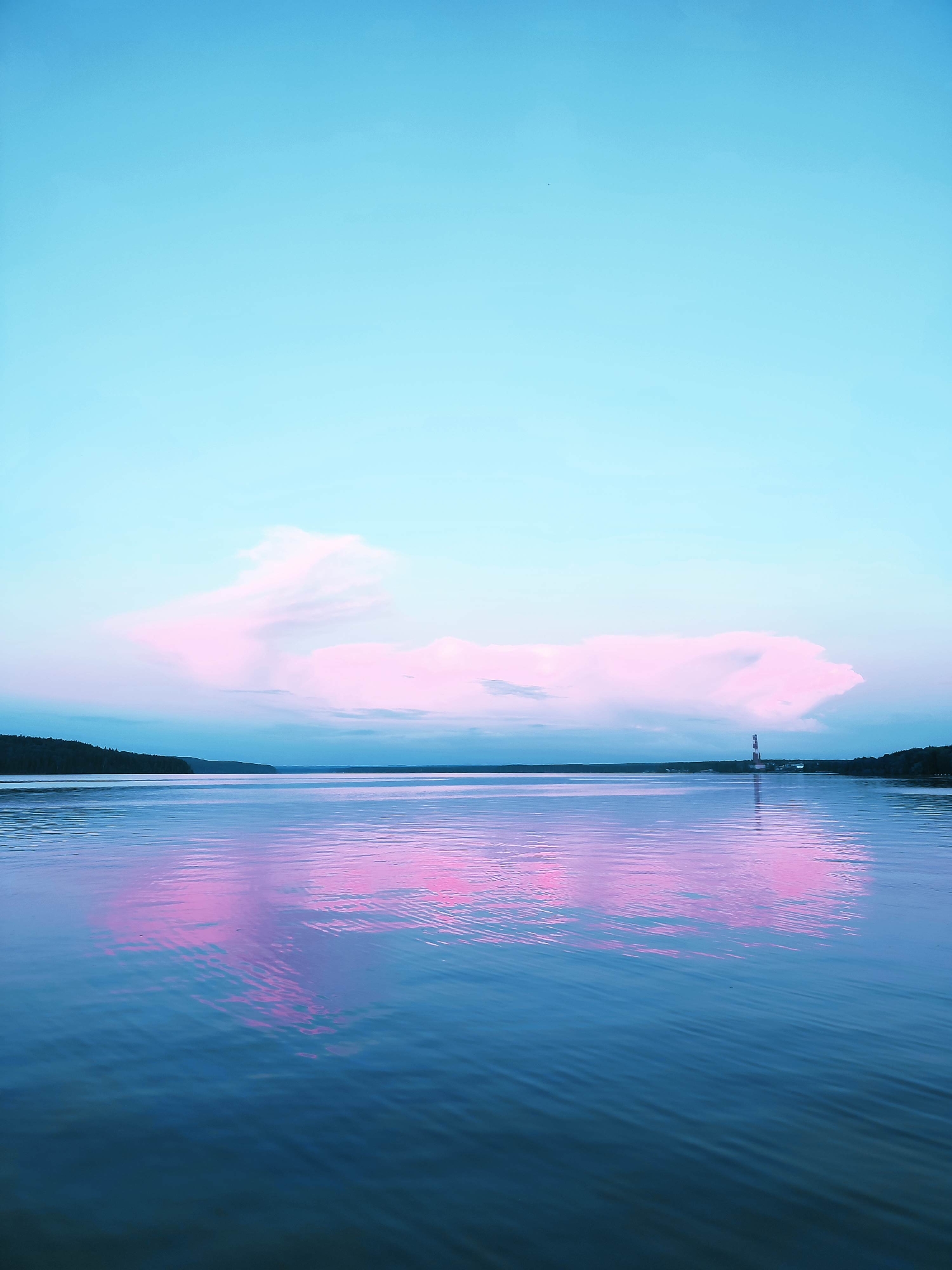 A wide shot captures a tranquil body of water under a serene, pale blue sky. The water's surface is a deep blue, reflecting the soft pink and white hues of a large, diffused cloud formation suspended in the sky. A thin line of dark green trees marks the distant horizon on the left side of the frame, and on the right, a slender, red and white striped structure, possibly a lighthouse or a navigational marker, stands against the horizon. The reflection of the cloud and the structure shimmers on the rippling water, creating an ethereal, dreamlike effect. The overall impression is one of peacefulness and natural beauty, with a hint of human presence in the form of the distant structure.