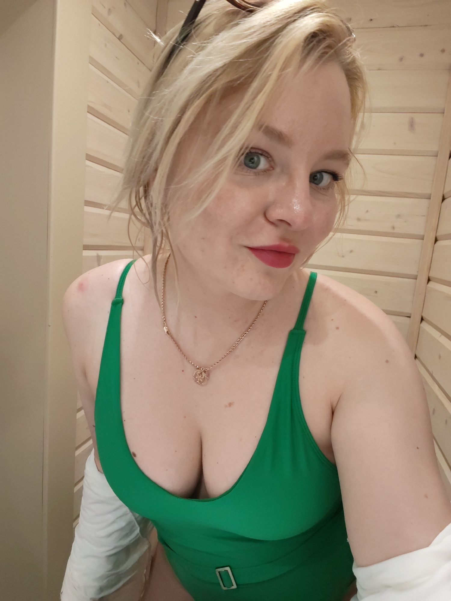 A fair-skinned woman with blond hair and blue eyes is posing for a selfie indoors. She is wearing a green one-piece swimsuit and a gold necklace with a round pendant. There are dark sunglasses perched on her head. A white towel or garment is visible on her left side. The background appears to be a wood-paneled wall. There is no information to suggest the photo was taken in Izhevsk, Russia.