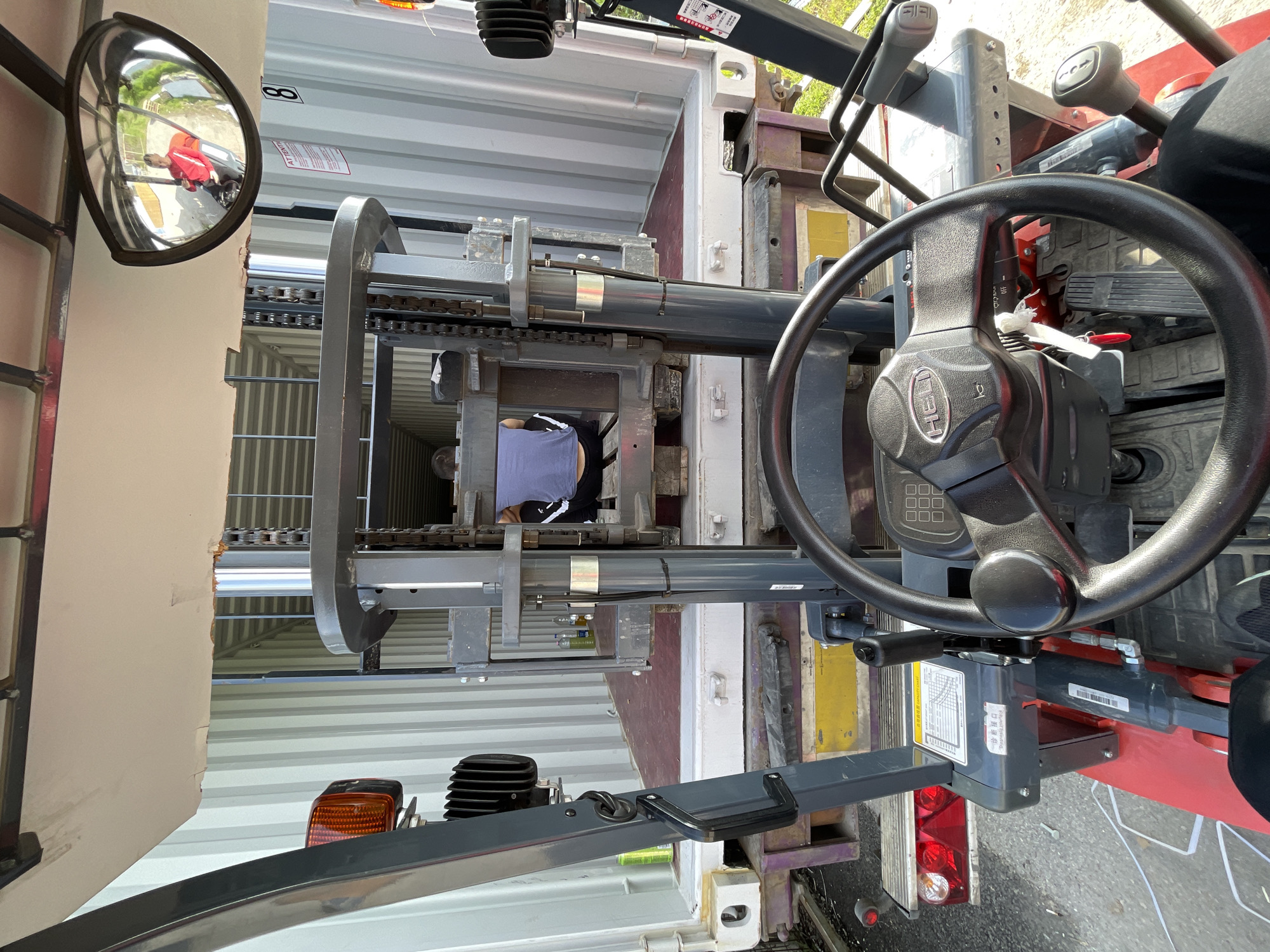 The image depicts the view from inside a forklift operating within a shipping container. The forklift is positioned inside the container, with the forks extended towards the back, where a person wearing a blue shirt and black shorts appears to be working or inspecting the load. The container's interior walls are ribbed and white.
The forklift operator's perspective includes the steering wheel, dashboard, and controls. A side-view mirror is visible on the left, reflecting a red object and possibly a vehicle outside the container.
The image does not provide sufficient information to confirm the location as Asenovgrad, Bulgaria.