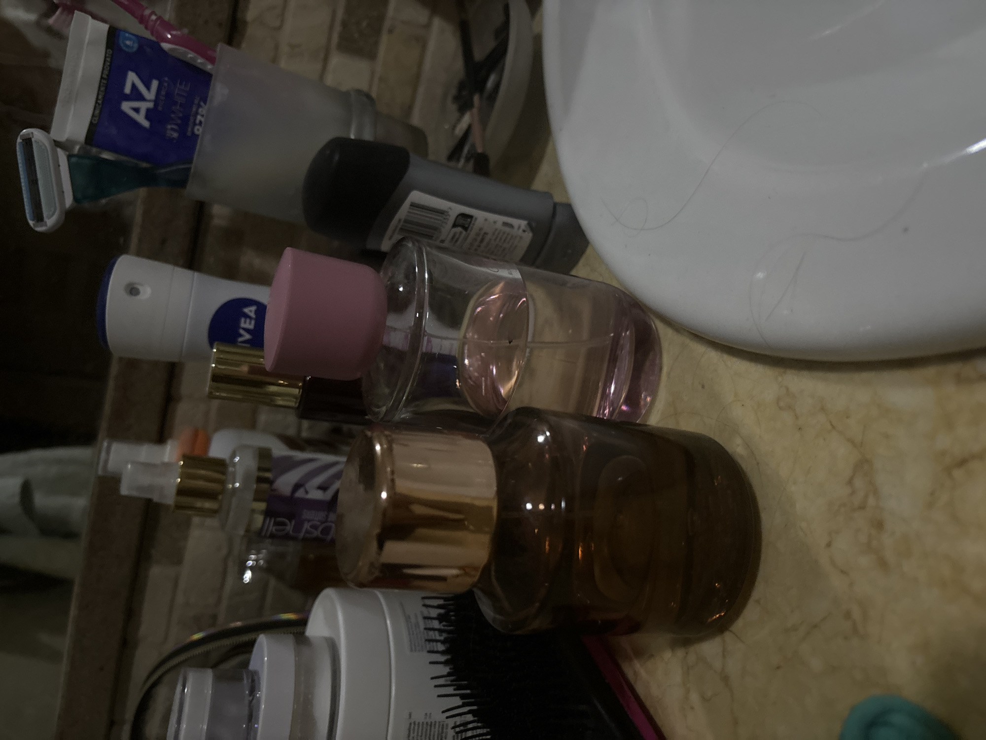 The image depicts a bathroom countertop cluttered with personal care items. A white sink is visible in the upper right corner. From top to bottom, the items include: toothpaste and toothbrush in a holder, a deodorant stick, a perfume bottle with a pink cap, a perfume spray bottle, a dark brown perfume bottle, and several other cosmetic bottles with a hairbrush. The surface appears to be a marble-like countertop. There is no evidence to suggest the location in Budva, Montenegro, or any people in the scene.