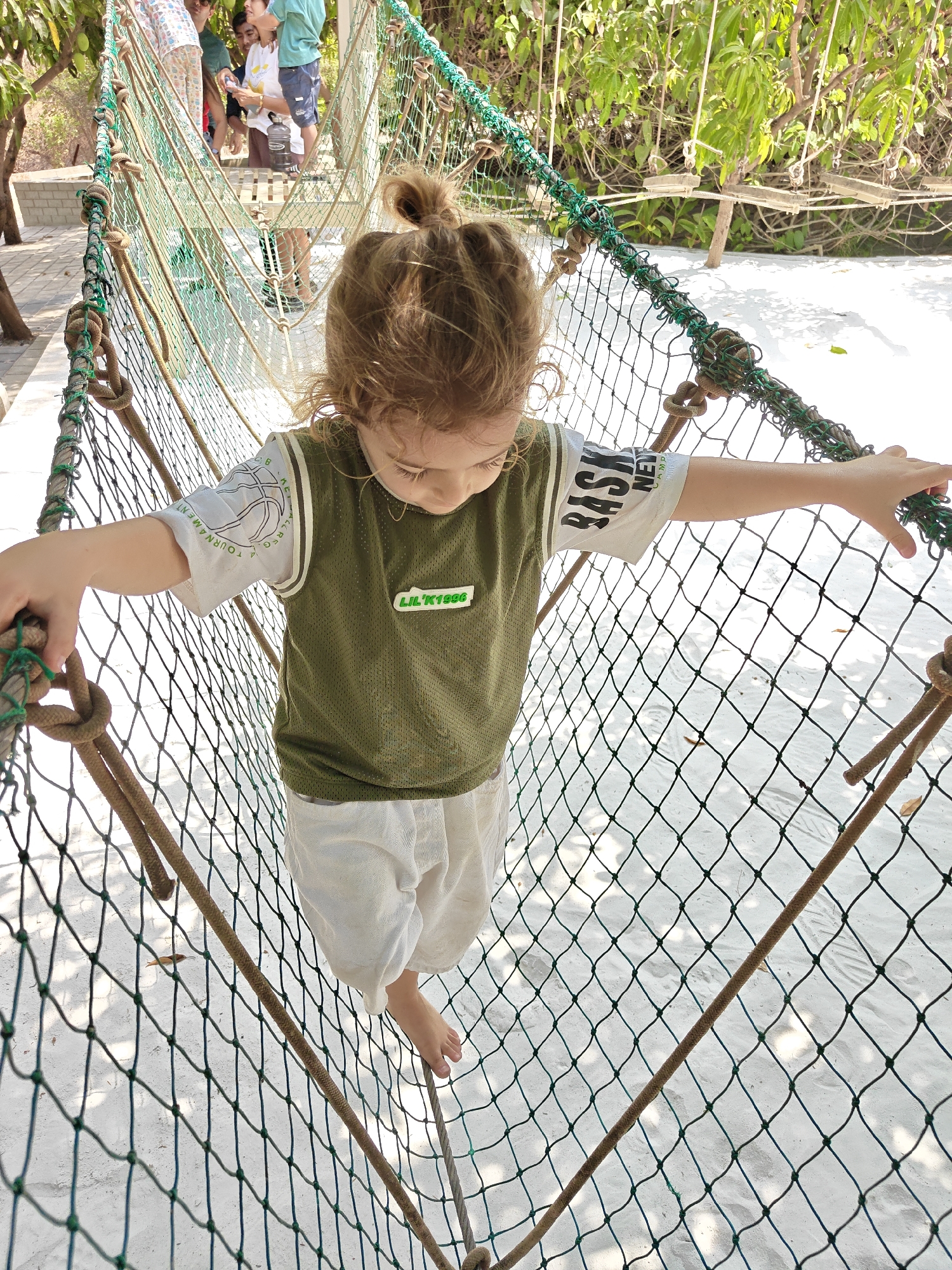 A young child walks on a rope bridge net at a play area in Kalote Mokashi, India. The child has light curly hair tied in a bun, wears a green jersey over a white t-shirt, and white shorts. They walk barefoot, using their hands to steady themselves on the ropes. The rope bridge is constructed of thick ropes and fine mesh netting. Other children and adults are visible in the background, standing on a platform connected to the bridge. Trees and foliage surround the play area. The ground beneath the bridge appears to be covered in light-colored sand.