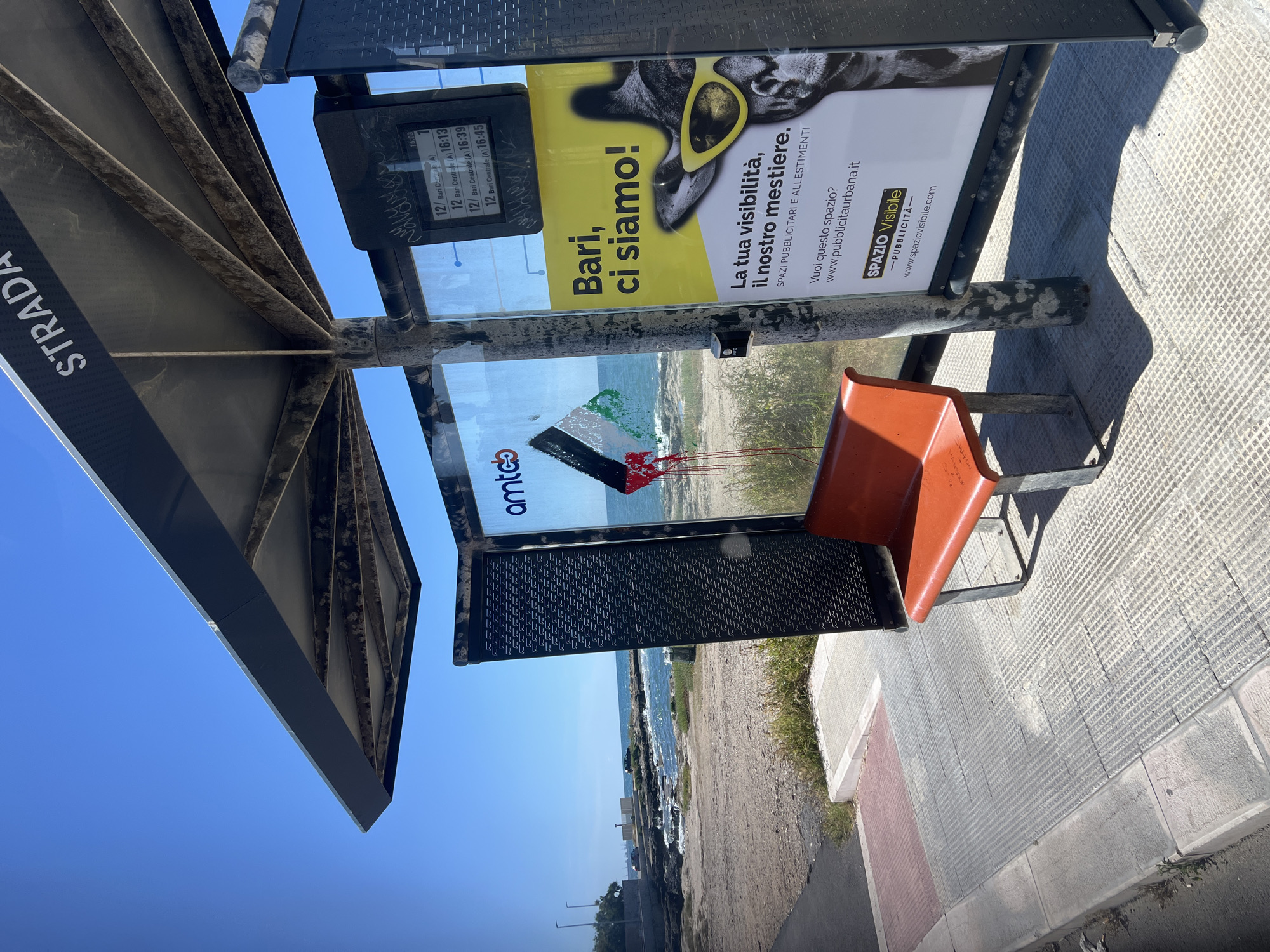 The image captures a bus stop situated by a coastline under a clear blue sky, likely in Bari, Italy. The setting appears bright and sunny, indicative of daytime.
The primary subject is a metallic bus shelter with a distinctive angled roof, displaying the word "STRADA" vertically on its side. Inside the shelter, an orange-red bench seat is visible. The shelter features glass panels, some of which are covered with advertisements. A digital display screen shows bus route information, indicating "16:03" as the current time, followed by bus line "12/ Bari Centrale (A)" with scheduled arrival times of "16:13", "16:39", and "16:45". Graffiti is scrawled on the glass panels beneath the display, including the word "HANSOUR".
One prominent advertisement features a stylized image of an animal, possibly a dog or deer, wearing yellow sunglasses against a bright yellow background. The ad copy, in Italian, reads: "Bari, ci siamo! La tua visibilità, il nostro mestiere. SPAZI PUBBLICITARI E ALLESTIMENTI" (Bari, here we are! Your visibility, our business. ADVERTISING SPACES AND FITTINGS). It also provides contact information: "Vuoi questo spazio? www.pubblicitaurbana.it". Another smaller ad below this reads "SPAZIO Visibile -PUBBLICITA- www.spaziovisibile.com". Partially visible on another panel is a logo reading "amtc" above an image that seems to depict a beach or sea scene with abstract colorful elements.
Beyond the bus stop, a narrow strip of land with sparse vegetation separates the pavement from the sea, which stretches out to the horizon under the bright blue sky. The pavement is textured, with lighter and darker sections, and appears to be made of concrete blocks. No people are visible in the scene, suggesting a quiet moment at the bus stop.