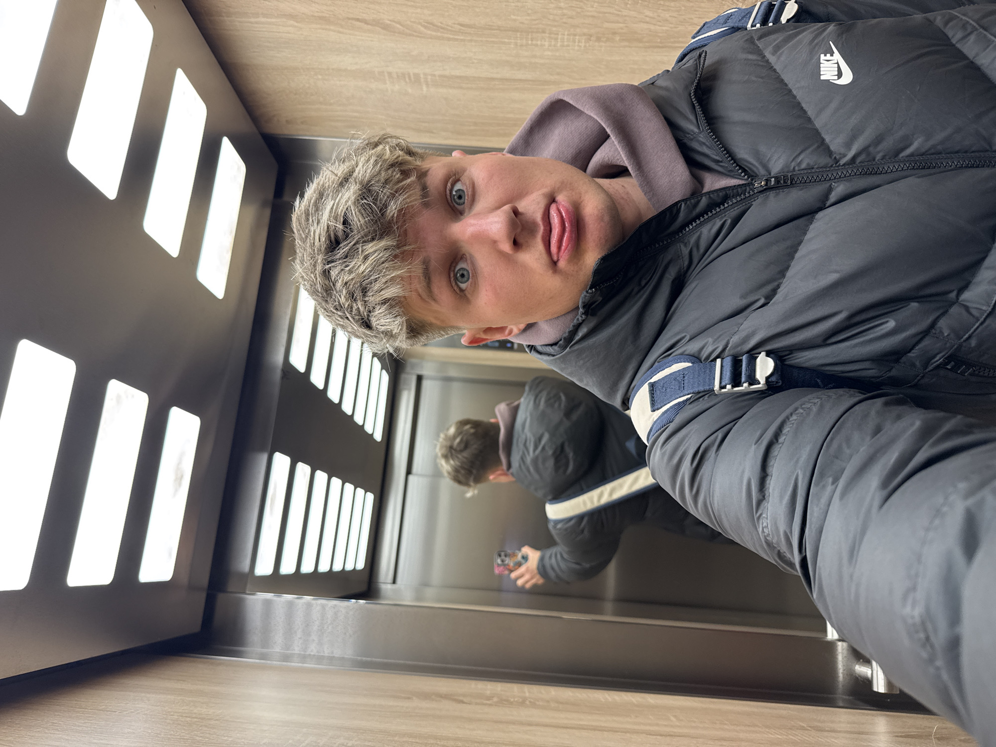 A young man is taking a selfie in an elevator. He is wearing a black Nike puffer jacket and a grey hooded sweatshirt. His hair is short and light-colored. He is sticking out his tongue and looking directly at the camera.
The elevator interior is modern with light wood paneling and a wall featuring evenly spaced, rectangular light cutouts. There is a mirrored wall reflecting the man and the rest of the elevator. The floor is also light-colored wood.
There is no information to determine that this scene is in the city of Plovdiv, Bulgaria.
