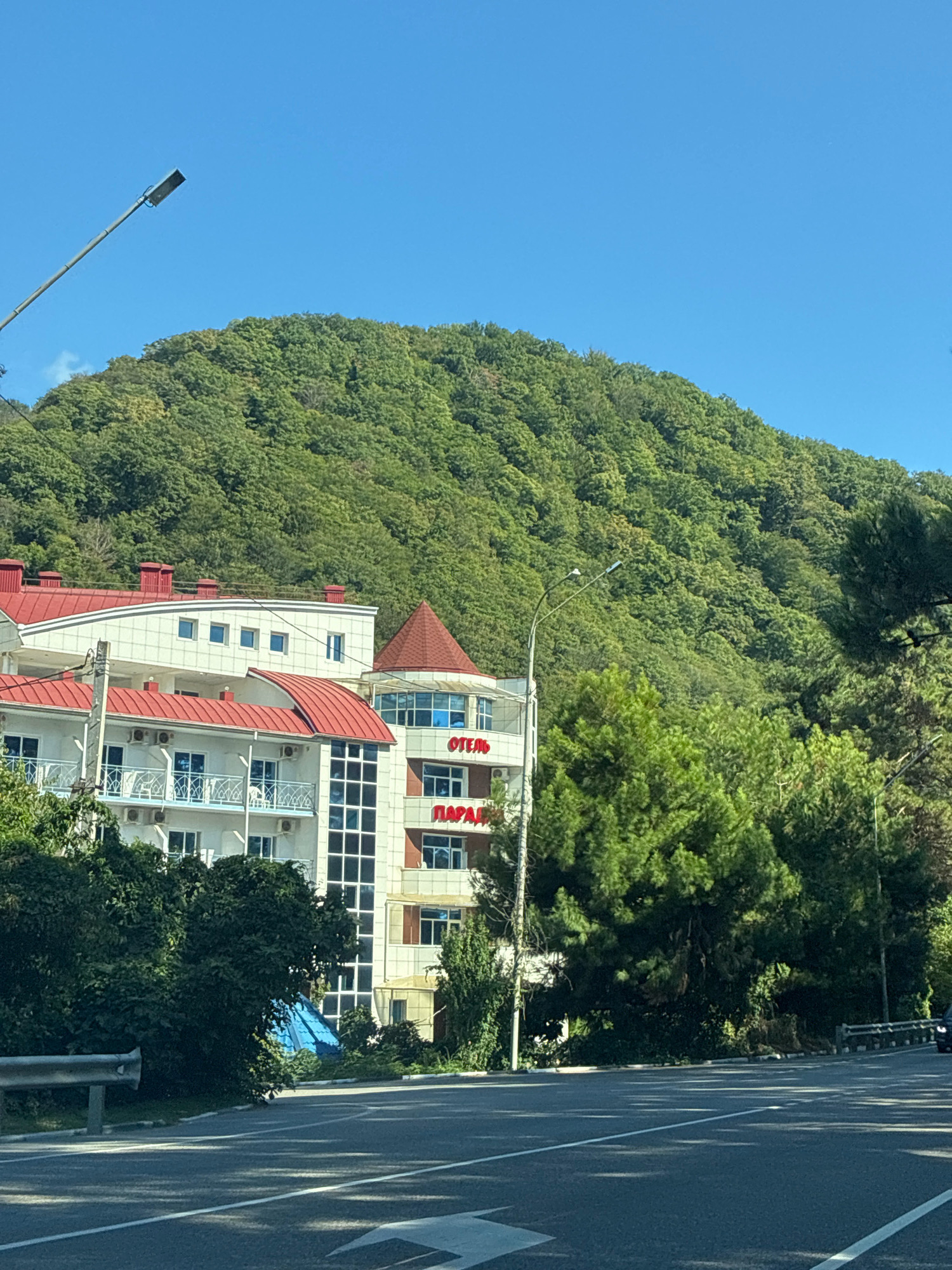 The image shows a white building with red roofs, identified by the Russian word "ОТЕЛЬ" (Hotel) and "ПАРАД" (Parade) in red lettering. The hotel is situated on a green, treed mountainside under a clear blue sky. A street runs in front of the hotel, with a guardrail visible. A street lamp is positioned on the left side of the image. The building appears to have multiple stories with balconies and windows. The surrounding environment is lush with trees, suggesting a natural, possibly resort-like setting in Olginka, Russia.