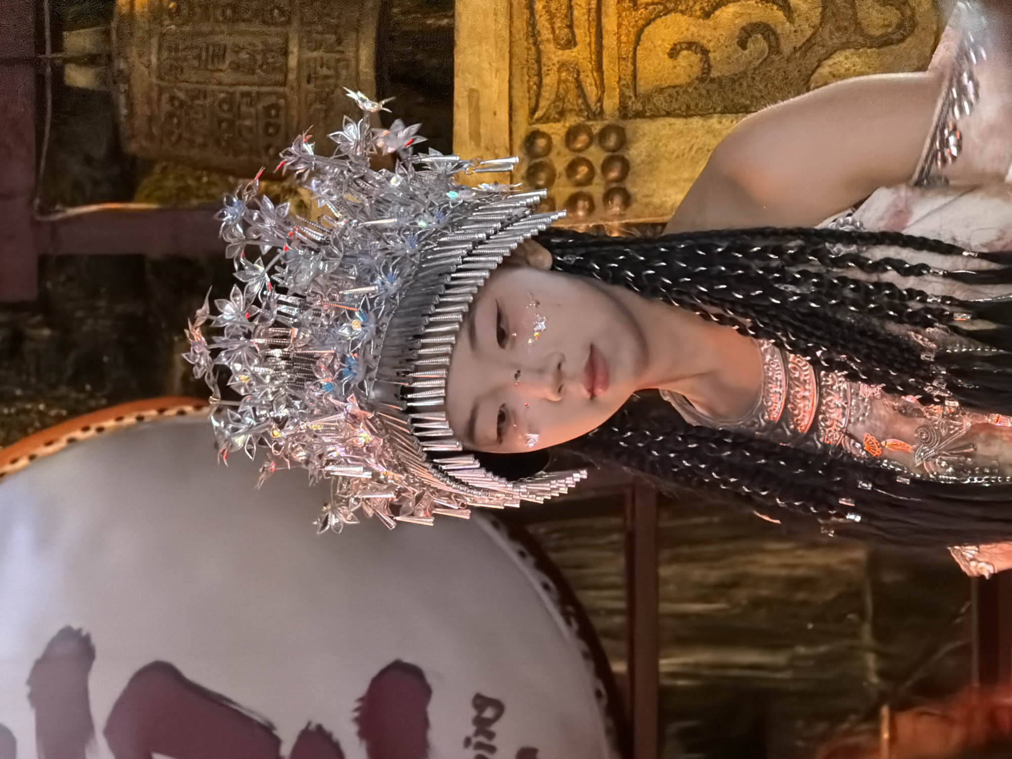 A young East Asian woman stands facing left in a headpiece adorned with silver metallic flowers and tassels. She has dark braided hair, some of which falls in front of her shoulder. Her face has makeup on it, including what appears to be glitter around her eyes and on her cheekbones. She wears a ornate silver outfit. In the background, to the left, is a large, round object with red Chinese characters on it, possibly a drum. To the right, there are decorative bronze-colored bells. The lighting in the scene is warm and golden, with a dark, possibly stage-like background. The location is the city of Yuzhong District, China.