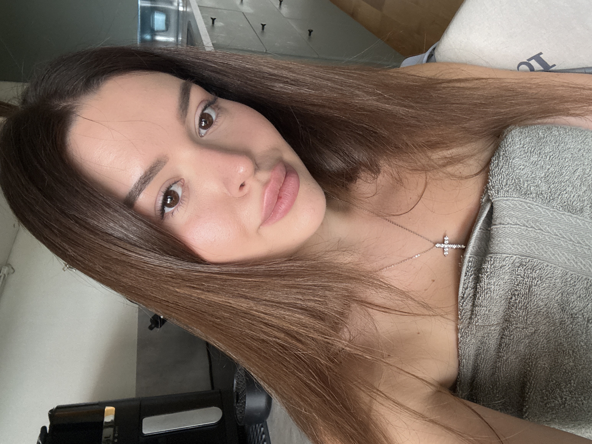The image shows a close-up selfie of a young woman with long, brown hair. She is wearing a grey towel around her chest and a silver cross necklace. Her skin appears to be glowing, suggesting she has recently showered or is wearing makeup. The background shows a kitchen setting with a black appliance and cabinets. The lighting is natural and soft, creating a relaxed atmosphere. The photo was likely taken indoors, in a residential setting.