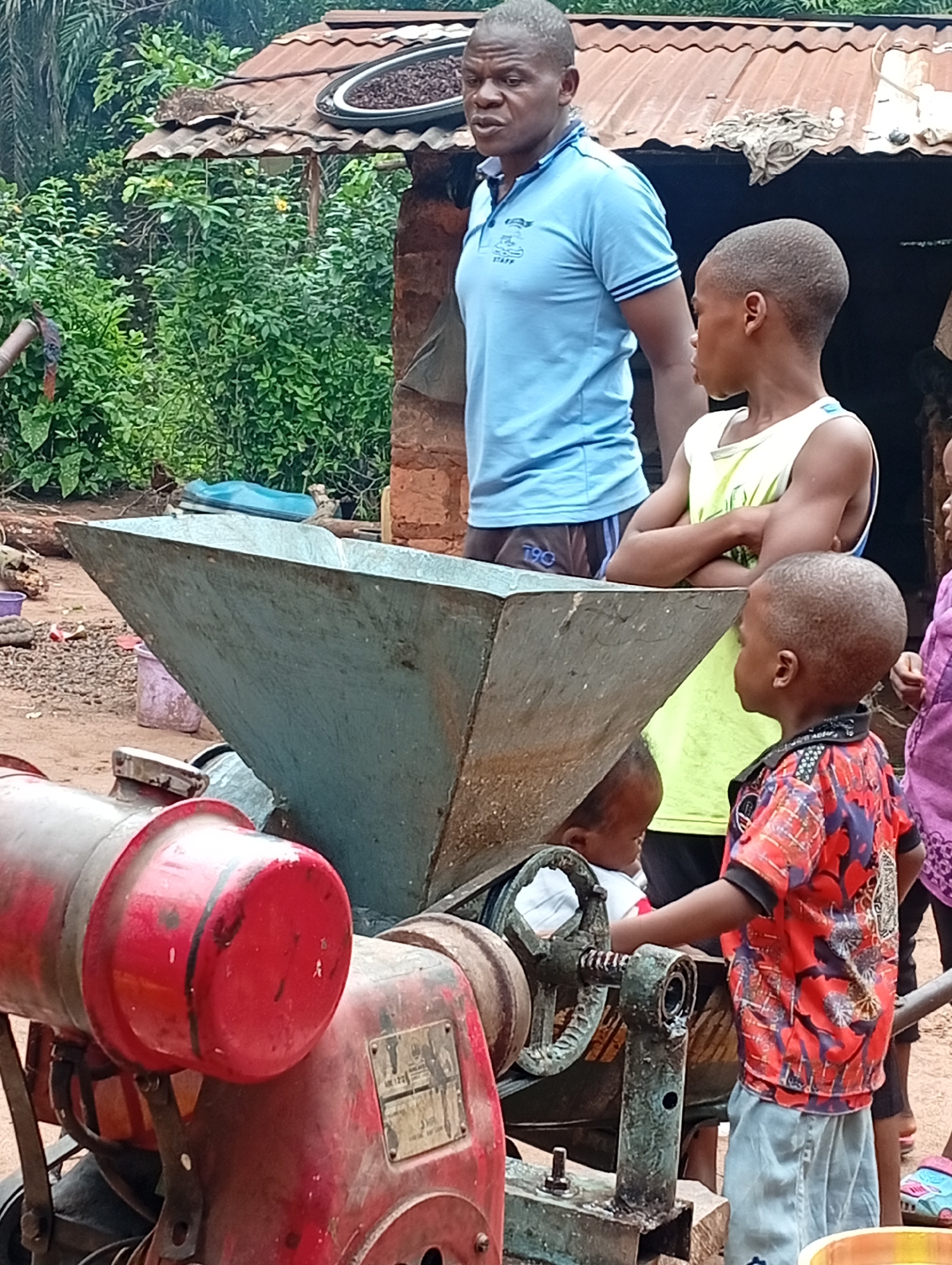 In Adda, Nigeria, a group of people, including children, are gathered around a mechanical device. A man in a blue polo shirt stands to the left, looking towards the right. To his right, a boy in a white tank top has his arms crossed, also looking to the right. In the foreground, a child in a red and black patterned shirt is reaching towards a part of the machine. Another young child is visible behind the machine. The machine itself appears to be a grinder or processing unit, with large metal components and a red engine on the left. The environment suggests a rural or semi-rural setting, with a rustic structure and lush green foliage in the background.