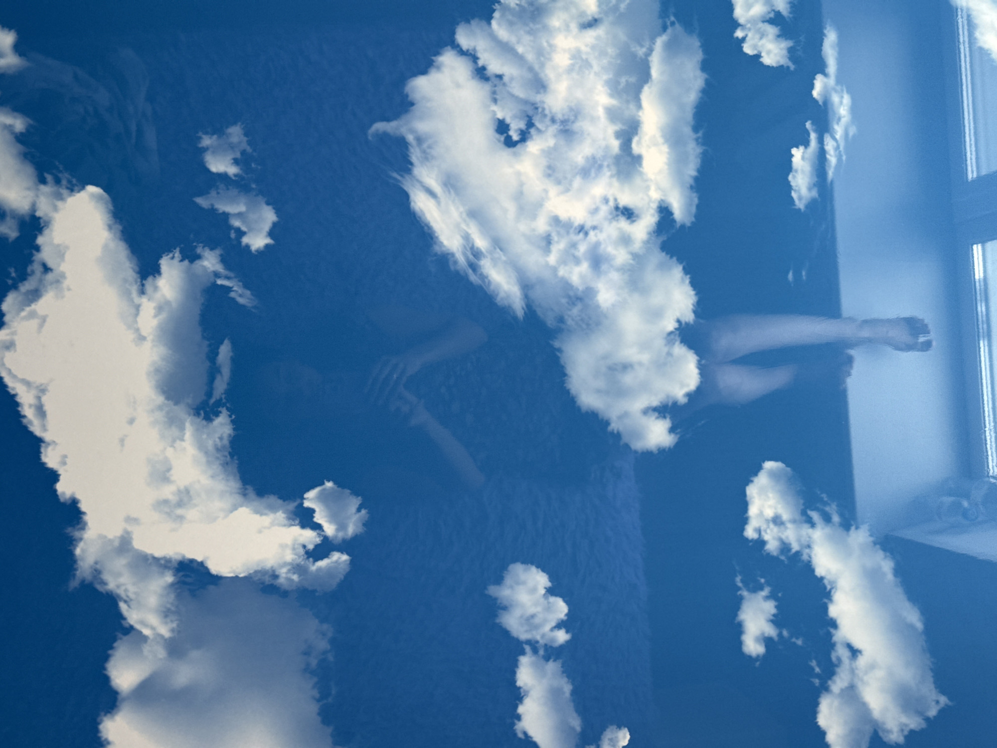 A surreal, dreamlike image combines an indoor scene with an expansive blue sky. A person is visible, lying on a dark blue or purple textured surface, possibly a bed or couch. Their form is partially obscured and translucent, suggesting a reflection, double exposure, or a filter effect. The individual appears to be resting or sleeping, with their arms seemingly crossed over their torso and legs slightly bent.
To the right of the person, a window with a visible frame allows light into the room and acts as a boundary. The entire scene is overlaid or reflected with a bright, clear blue sky filled with numerous fluffy white clouds, giving the impression of looking up at the sky, or having the sky reflected onto the interior space. The lighting suggests it is daytime with good weather. The combination of the resting figure and the vast sky creates a tranquil and contemplative atmosphere, blending indoor comfort with the openness of nature. No text is visible in the image.