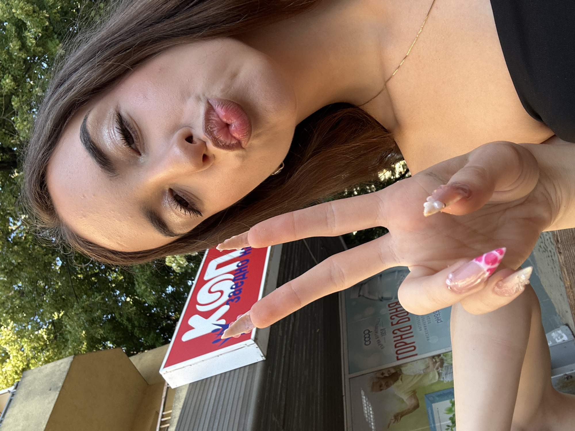 A young woman with long brown hair and striking makeup, including glitter eyeshadow and dramatic mascara, is taking a selfie outdoors. She is puckering her lips and holding up two fingers in a peace sign. Her fingernails are long and elaborately decorated with pink and white designs and small pearls. She is wearing a black strapless top and a delicate gold necklace. In the background, a building with a red sign that reads "КОП" and Cyrillic text is visible. Further behind, a poster for "COOP Горна Баня" with images of elderly people can be seen. The overall setting appears to be an urban environment with trees and foliage suggesting a public space in the city of Shumen, Bulgaria.