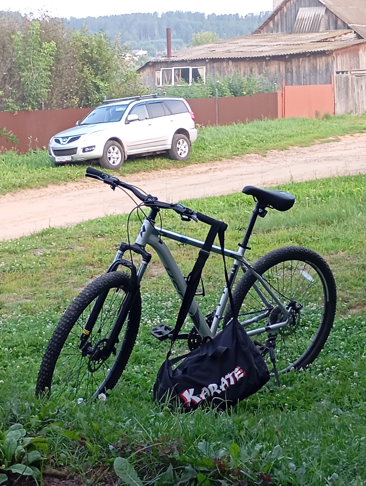 A bicycle with a black bag attached is parked on the grass in front of a dirt road. The bicycle is silver and black with black tires and pedals. The bag is black with "KARATE" written in red and white letters. In the background, a white SUV is parked on the dirt road. Behind the SUV is a brown fence, and further back is a weathered wooden building with a corrugated metal roof. The scene appears to be in a rural or semi-rural area, possibly a village or the outskirts of a town, given the presence of a residential-style building and natural landscaping. The overall environment suggests an outdoor setting with vegetation and a dirt road, likely in a temperate climate based on the green grass and trees. The location is identified as Fokino, Russia, indicating a specific geographical context for the scene. There are no people visible in the image.