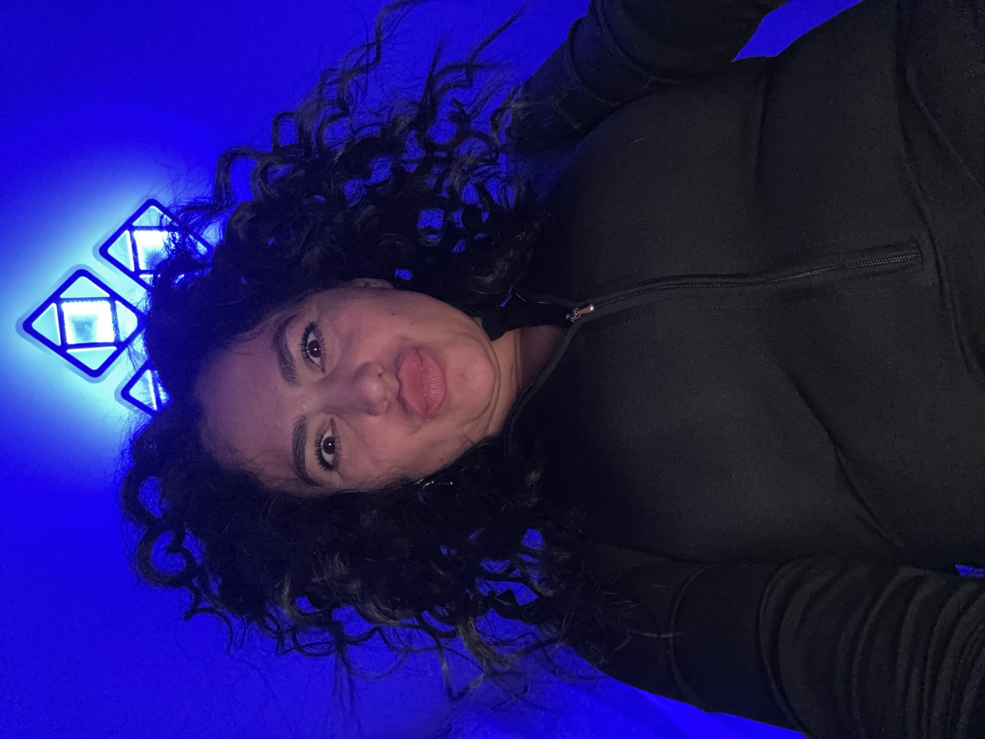 A woman with curly dark hair is posing indoors. She is wearing a black zip-up jacket. Behind and to the left of her is a blue illuminated wall with two geometric light fixtures. There is no evidence to indicate a location in Kauchuk Stantsiya, Russia.