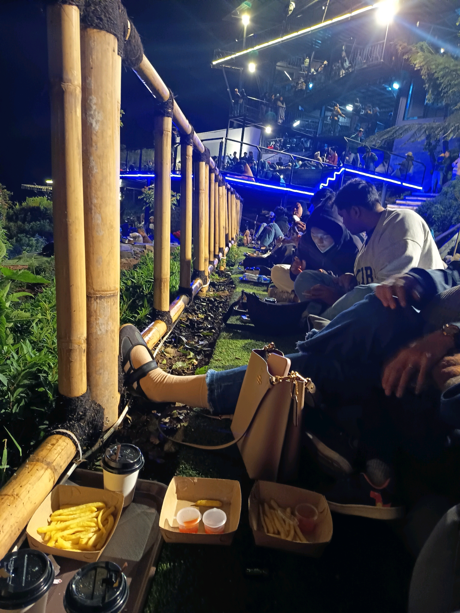The image depicts an outdoor nighttime scene at a multi-level establishment in Keditan, Indonesia, likely a cafe or restaurant, with numerous patrons. A bamboo railing, bound with dark rope, runs along the foreground, separating the viewer from a grassy area where several individuals are seated on the ground. In the foreground, a tray holds french fries and two small cups of dipping sauce, alongside a disposable coffee cup. A woman's legs, clad in tan stockings and sandals, are visible resting on the ground, with a tan handbag nearby. Further back, on the lower and upper levels of the establishment, groups of people are seen mingling and sitting, illuminated by bright blue and white lights. The overall atmosphere appears to be one of casual social gathering.