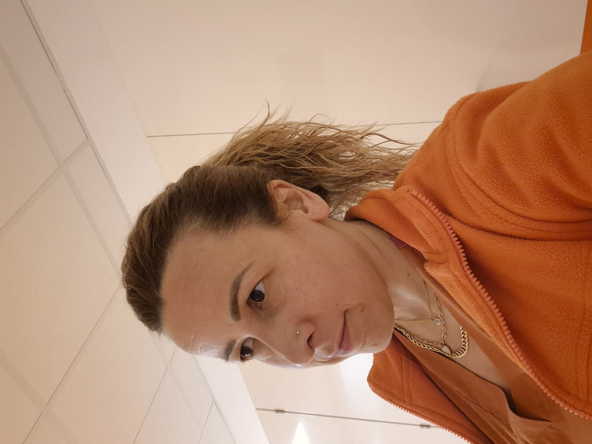A woman with brown hair tied back in a partial ponytail and wearing an orange fleece jacket is indoors, likely in a public restroom. She is also wearing necklaces and has a nose piercing. The background consists of white wall panels. The scene is tilted. The location is Unknown, Unknown.