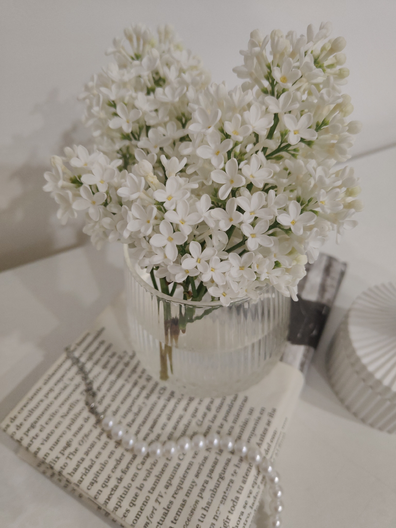 A close-up, slightly high-angle shot features a delicate arrangement of white lilacs in a clear, ridged glass vase. The vase sits atop a stack of open books with printed text in Spanish, partially obscured by a string of faux pearls. Next to the book, a small, round, decorative box with a pleated design sits on the surface. The background is a plain, light-colored surface, possibly a table or countertop, suggesting an indoor setting. The overall color palette is soft and muted, with a focus on whites and creams.