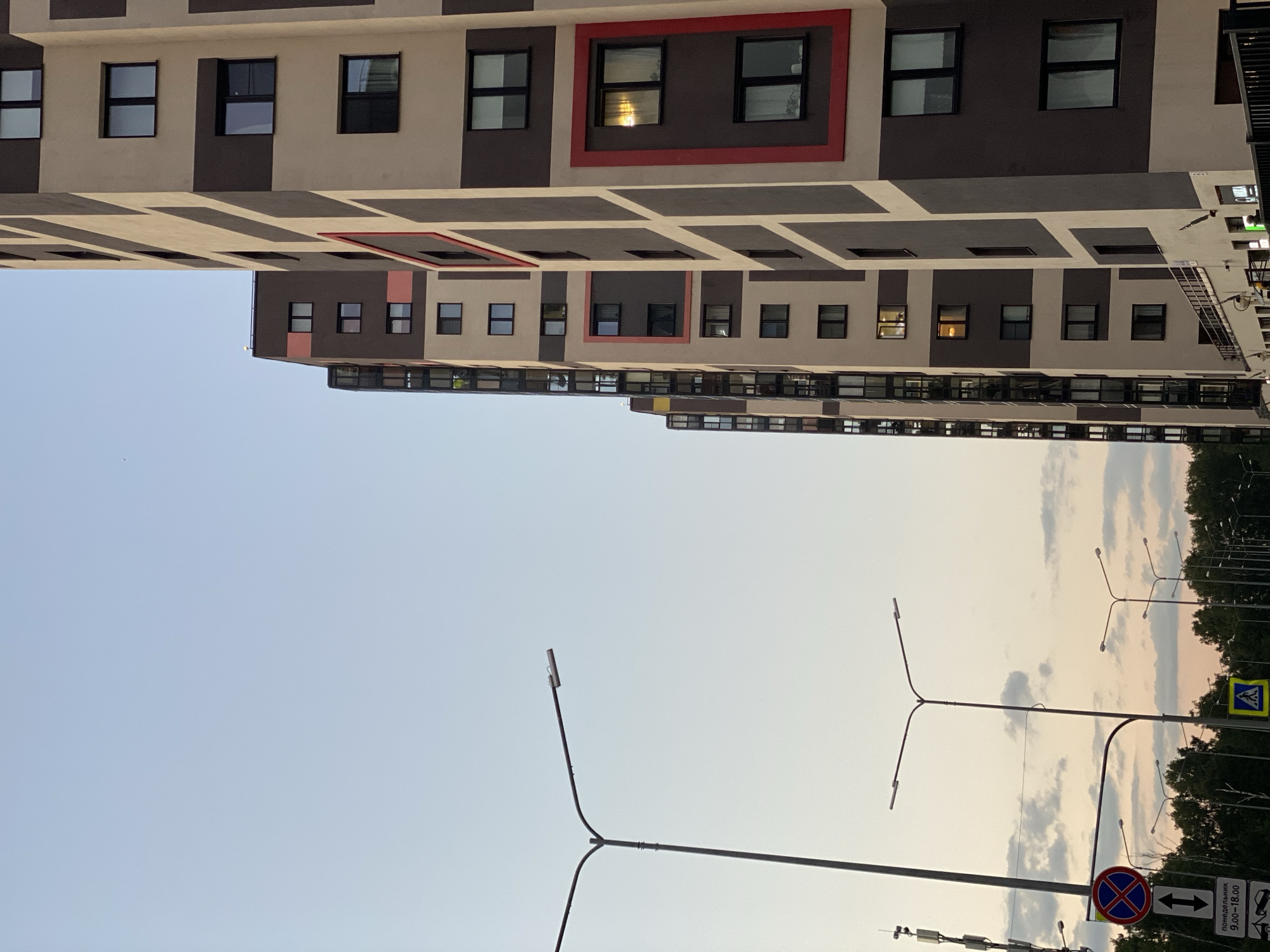 The image captures a cityscape scene in Kudrovo, Russia, featuring modern residential buildings against a pale blue sky. The primary focus is on a row of tall, multi-story apartment buildings with a distinctive color scheme of beige and dark brown vertical stripes. Some buildings have red accents around certain windows. Numerous dark-framed windows are visible, with some illuminated from within, indicating occupancy.
In the foreground, several streetlights stand tall, one prominently on the left with a Y-shaped pole and another in the middle ground with a similar structure. Traffic signs are also visible: a circular red and blue sign with a white horizontal bar and a white vertical arrow, along with a rectangular white sign below it displaying text and numbers in black, suggesting parking or time restrictions. A yellow pedestrian crossing sign is also present.
A line of lush green trees forms a backdrop at the bottom of the frame, separating the buildings from the lower ground. The lighting suggests it is either early morning or late evening, with a soft, diffused light illuminating the scene. There are no people or vehicles immediately apparent in the frame. The overall impression is one of urban residential development.