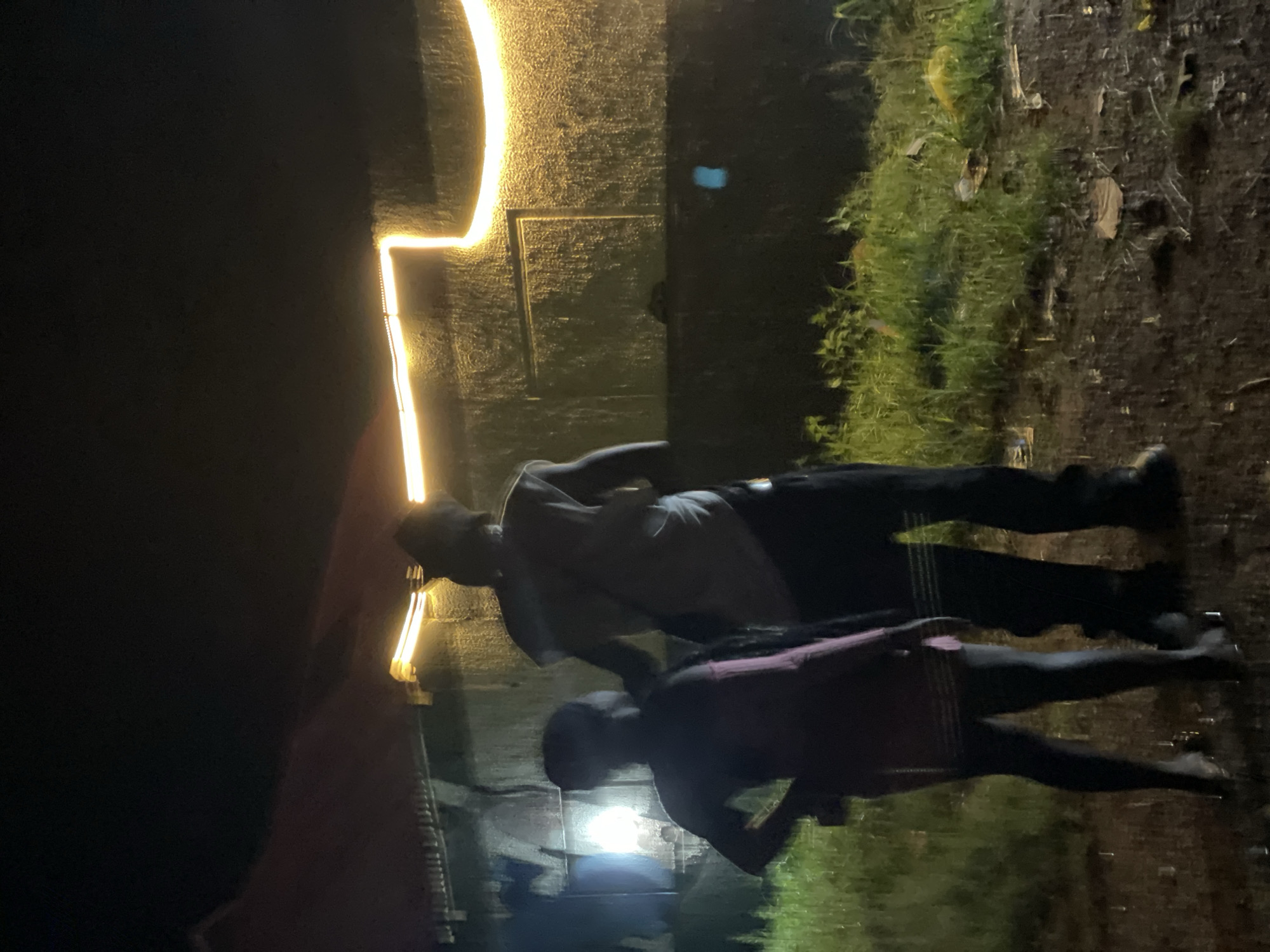 Two figures stand in front of a building at night in Durumi, Nigeria. The figure on the left is smaller, appearing to be a child, wearing a pinkish garment. The figure on the right is taller, possibly an adult, wearing a light-colored shirt and dark pants. They are silhouetted against the building, which is illuminated by a horizontal strip of bright yellow lights. The ground in front of the building is wet and appears to be a mix of dirt and grass, with some scattered debris. The overall atmosphere is dim, suggesting nighttime conditions.