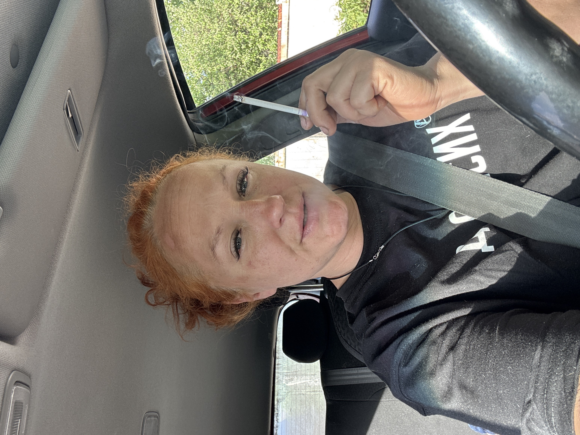 A woman with auburn hair sits in the driver's seat of a car, wearing a black t-shirt and a seatbelt. She is holding a lit cigarette. The location is identified as Chaikovsky, Russia based on the shirt. The interior of the car is visible. Outside the car window, greenery and a building can be seen.