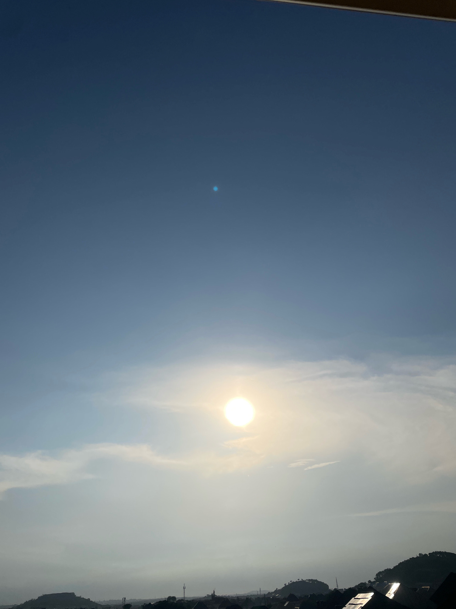This image captures a panoramic view of the sky over Abuja, Nigeria, at dusk. The sun, a bright, orb of light, is positioned slightly left of center in the frame, partially obscured by thin, wispy clouds. The sky transitions from a deep blue at the top of the image to a lighter, hazy blue near the horizon. A small, distinct blue speck, possibly a lens flare or a distant object, is visible in the upper portion of the sky.
Below the sky, the silhouette of the city of Abuja is discernible. The foreground is dominated by the dark shapes of rooftops, some reflecting the fading sunlight with a bright sheen. Hills, covered in dark foliage, rise gently in the mid-ground, dotted with the outlines of buildings and telecommunication towers. The overall impression is one of a serene evening cityscape, with the natural beauty of the sky contrasting with the man-made structures below. The image appears to have been taken from an elevated vantage point, possibly a building or a hill overlooking the city.