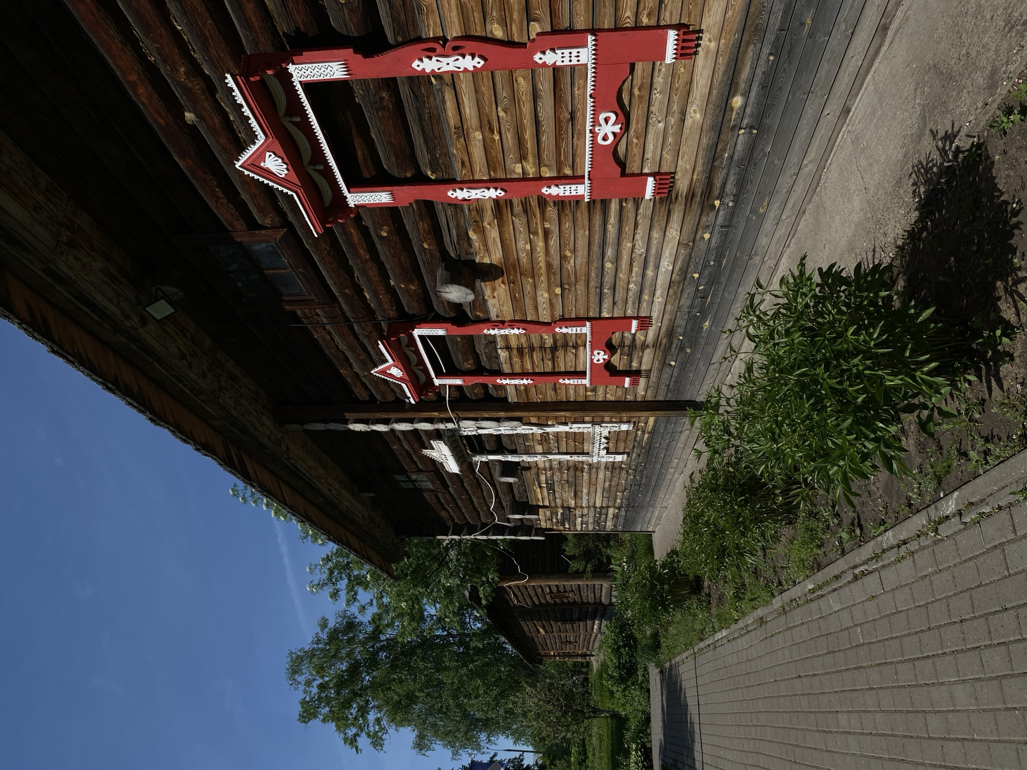 The image captures a scene in Votkinsk, Russia, featuring traditional wooden architecture. A long, low wooden structure with intricate red and white trim along the roofline dominates the upper portion of the image. The building's log walls are visible, with a window and a light fixture partially visible on the left side. A paved walkway runs parallel to the building, separated by a narrow strip of soil containing plants. The sky is a clear blue in the lower left corner, and trees frame the image. Another wooden building is visible further down the walkway, suggesting a row of similar structures. The overall impression is of a historical or cultural site showcasing Russian vernacular architecture.