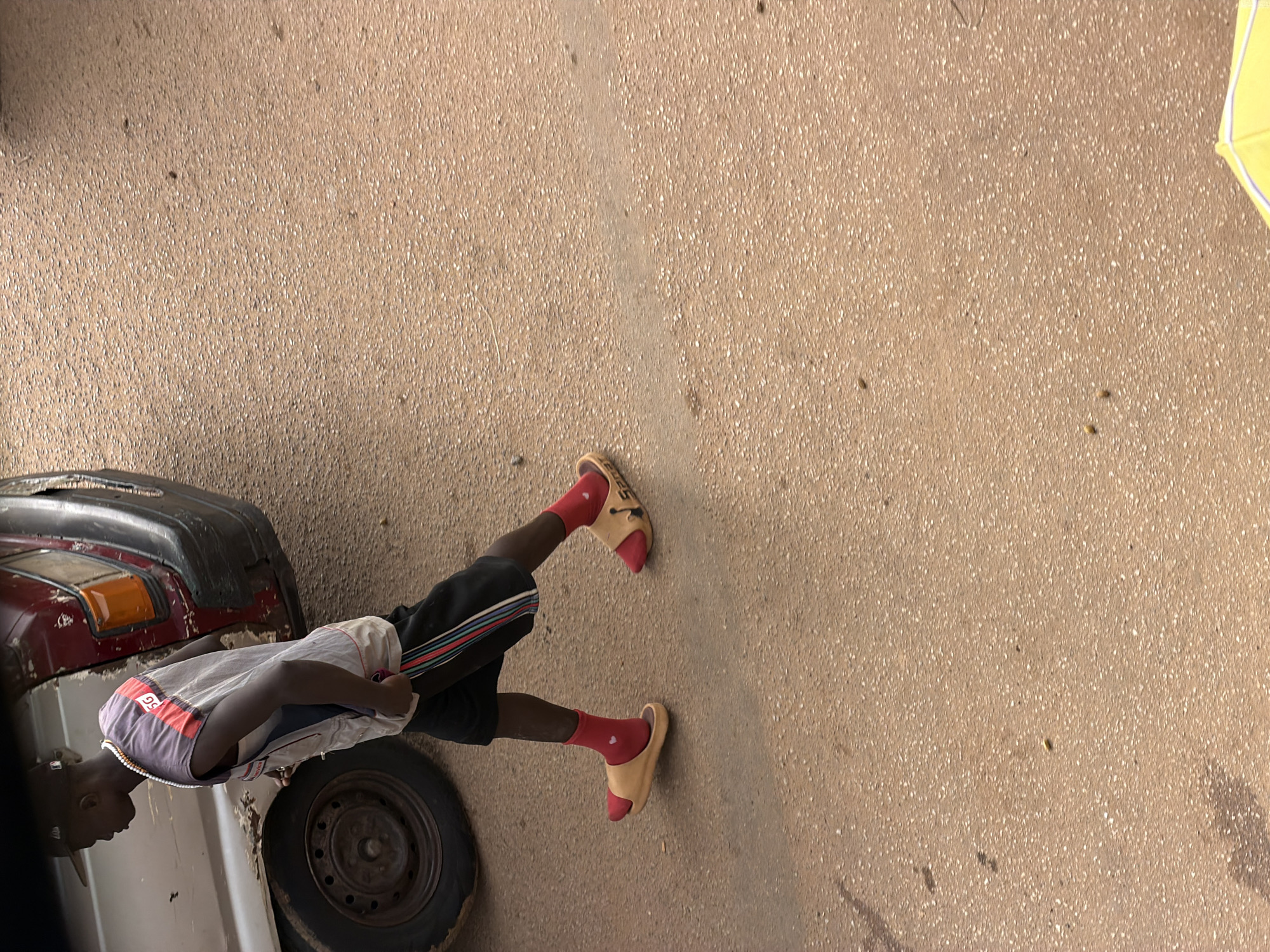 The image shows a person walking on a dirt road in Bukuru, Nigeria. The person is wearing a sleeveless shirt, black shorts with red and green stripes, red socks, and tan slides. A portion of a red vehicle is visible on the left side of the image. The road surface is reddish-brown and speckled with small white particles. The lighting suggests it is daytime.