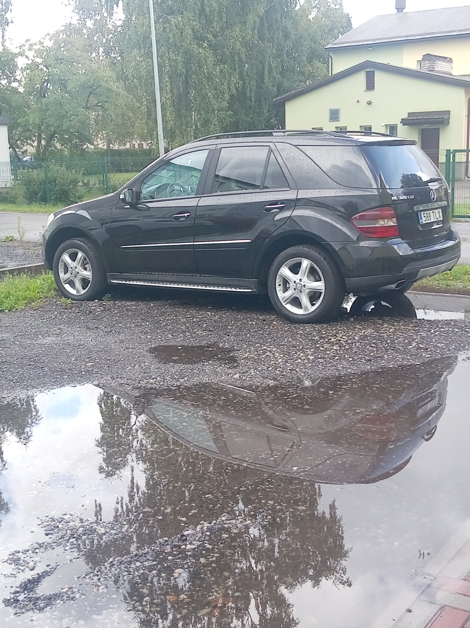 A black Mercedes-Benz ML 320 CDI SUV is parked on a gravel surface next to a large puddle. The car is facing to the right, and its reflection is visible in the puddle. The background shows a light green building with a dark roof, trees, and a white utility pole. The weather appears to be overcast, as indicated by the reflection of the sky in the puddle. The license plate of the car reads "588 TLX".
